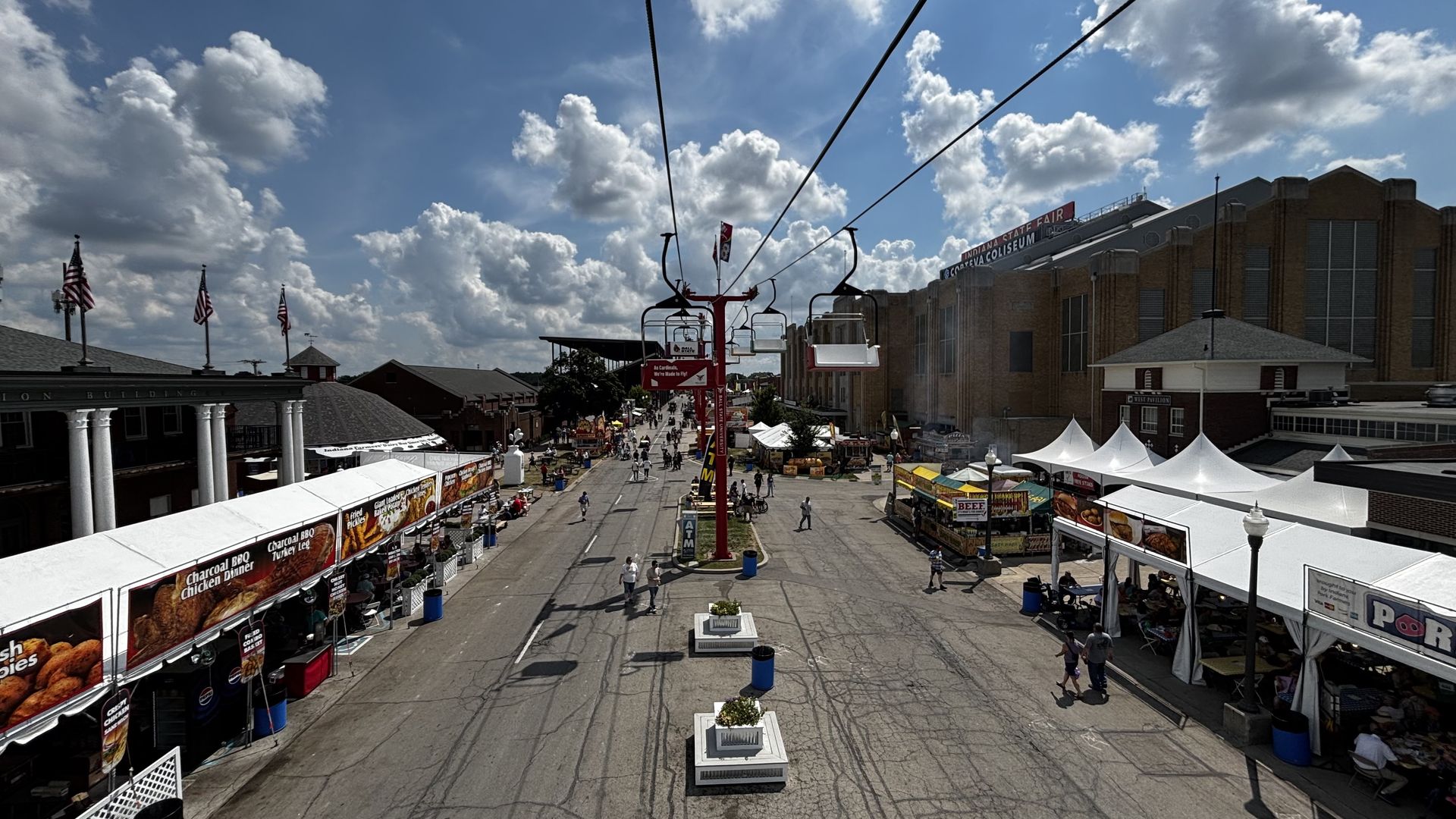 Aerial view of Indiana State Fair with food stalls on both sides of the street, a red chairlift going overhead, blue sky with clouds, and people walking along the fairground.