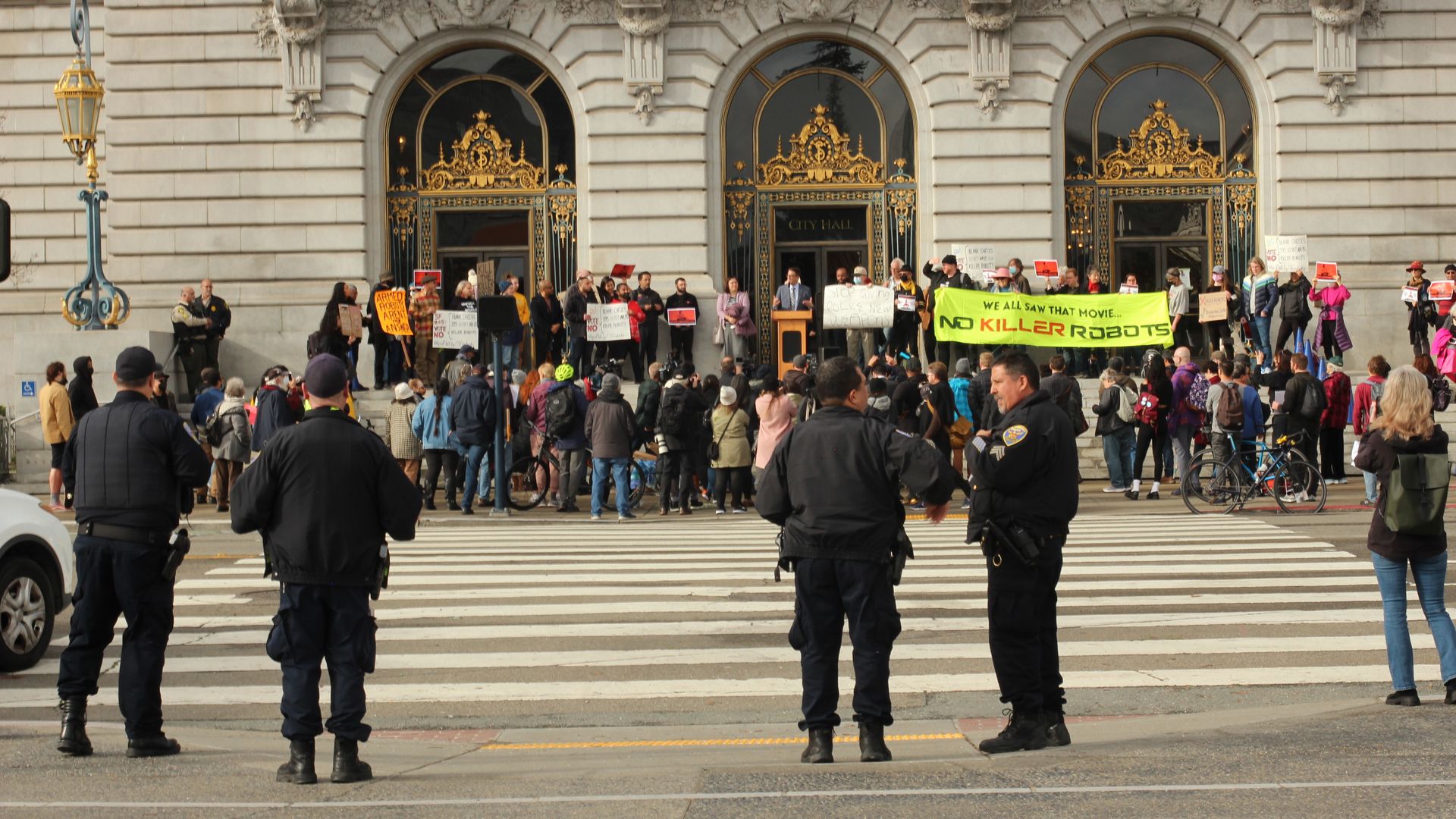 Police officers standing in front of rally outside city hall where protesters are holding sign that says "No killer robots"