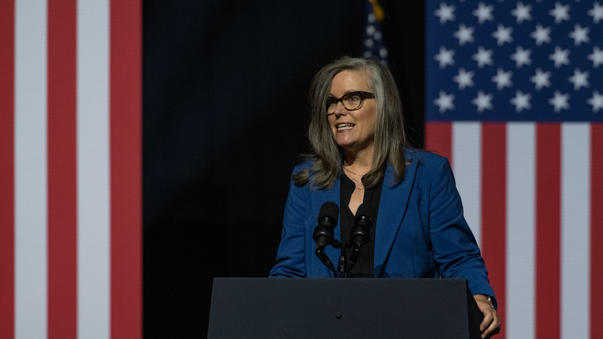 A woman in a blue suit with a grey streak in her hair speaking between two American flags. 