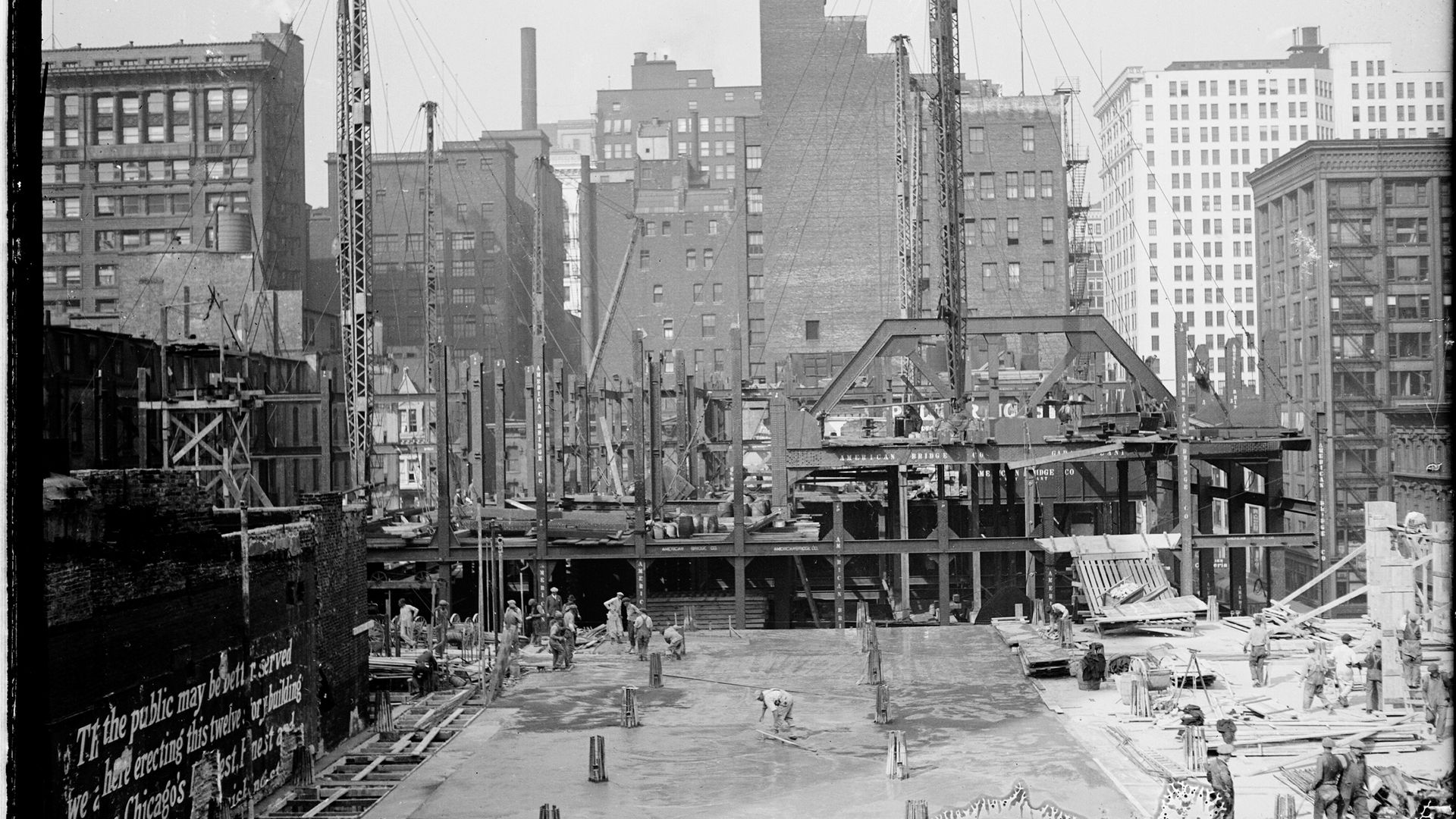 Black-and-white photo of a city construction site: workers on ground amid steel scaffolds and tall cranes; brick buildings and smokestacks loom in the background.