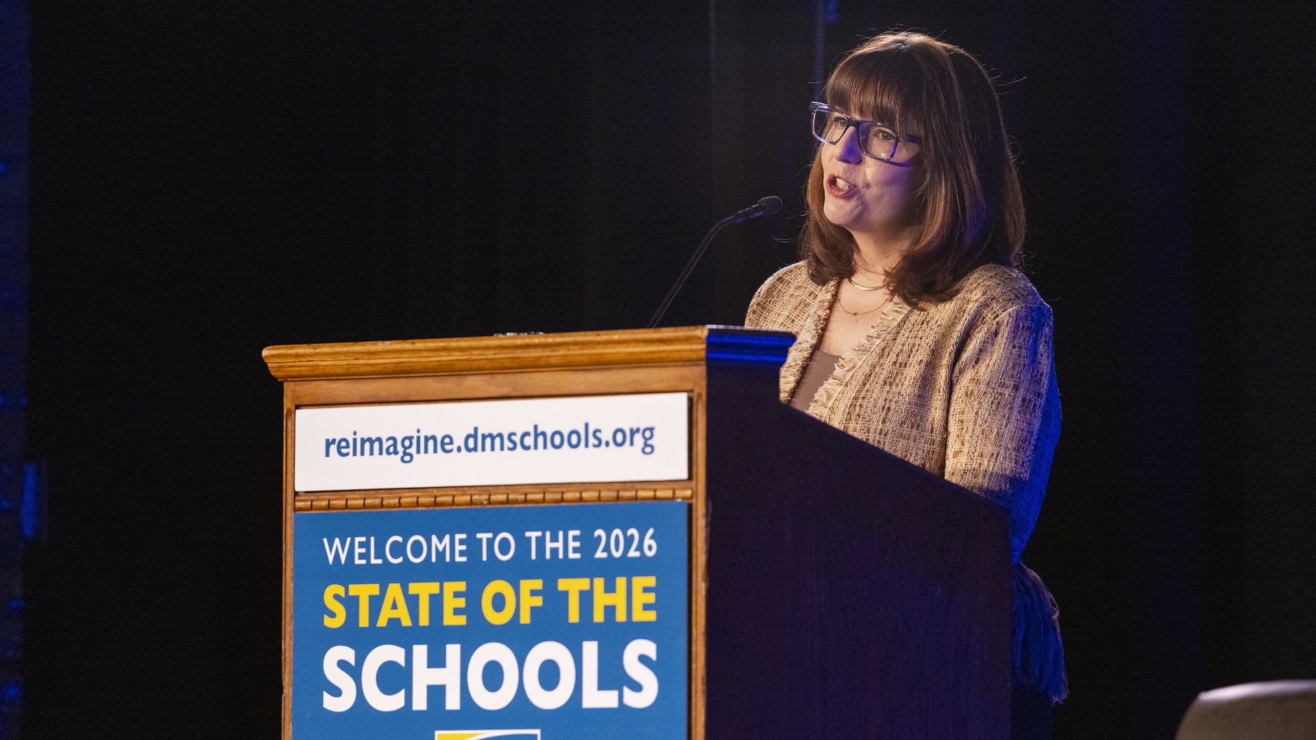 Woman with glasses speaks at a wooden podium; banner reads "Welcome to the 2026 State of the Schools" with the Des Moines Public Schools logo and reimagine.dmschools.org.