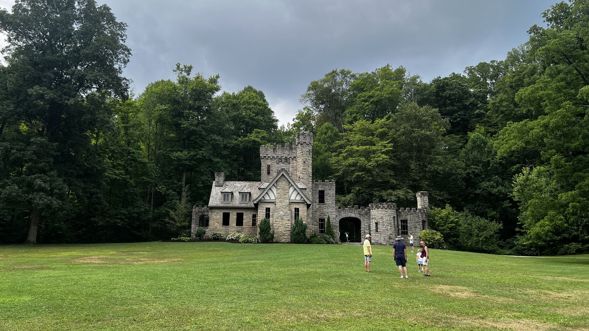 Stone castle-like building with battlements rises from a green lawn, framed by tall trees. Overcast, gray clouds fill the sky as a small group of people stands by the arched entrance.