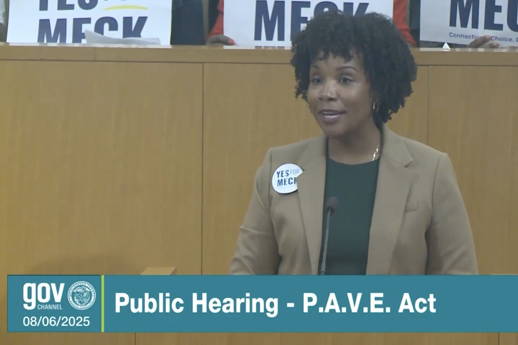 A woman with curly hair wearing a tan blazer and green top speaks at a public hearing on the P.A.V.E. Act, wearing a "YES FOR MECK" button; signs with "YES FOR MECK" are visible behind.