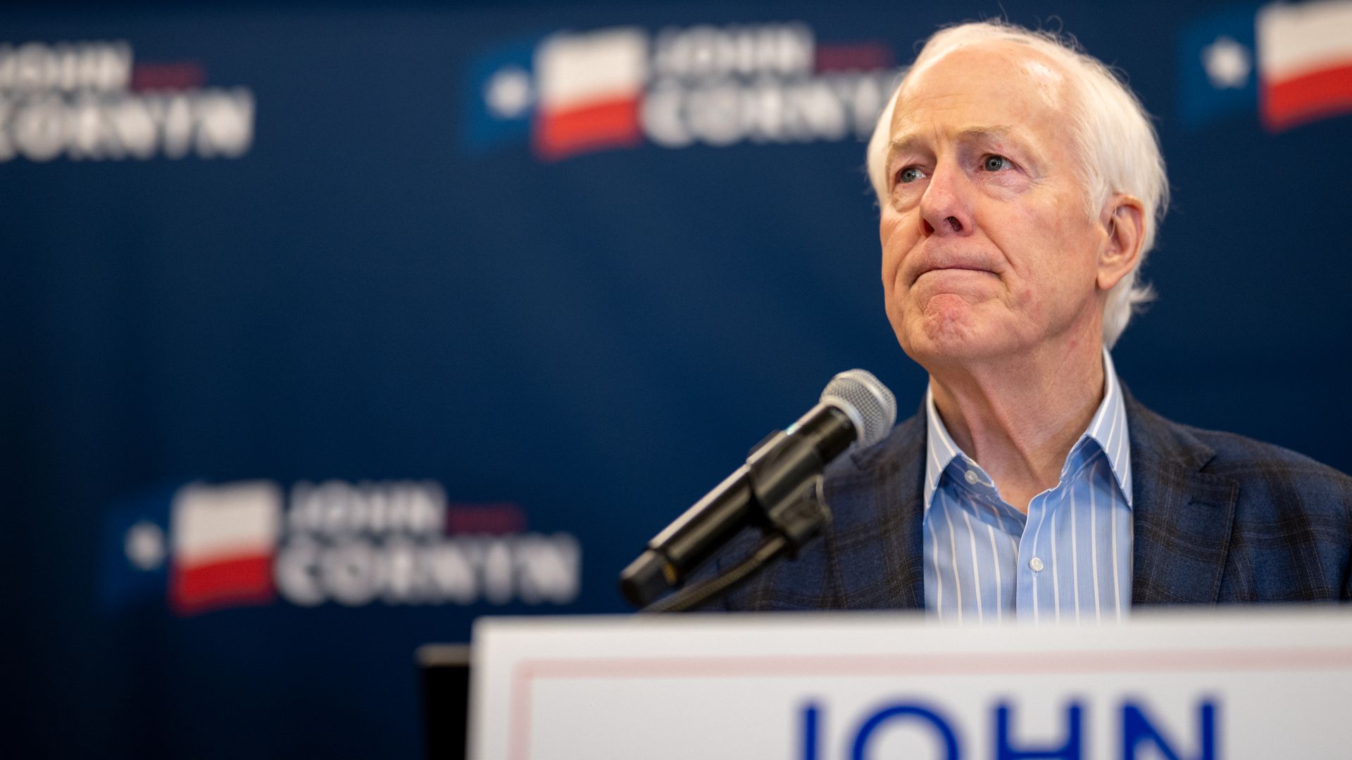 U.S. Sen. John Cornyn (R-Texas) in front of a microphone on a podium, with campaign imagery blurred in front of the podium and behind him. His face is stern and reflective.