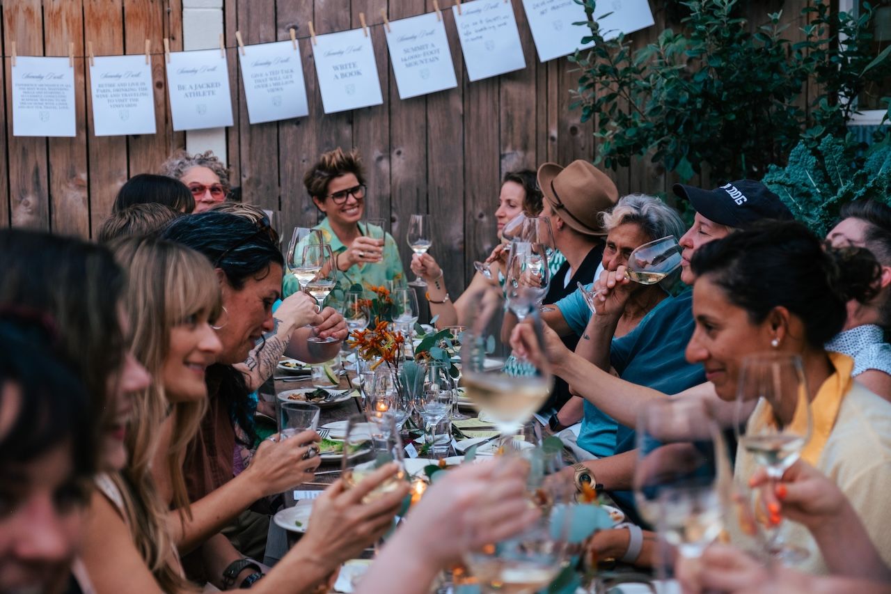 People enjoying drinks and conversation at an outdoor gathering, seated at a long table decorated with flowers, against a wooden backdrop displaying event cards, capturing community camaraderie.