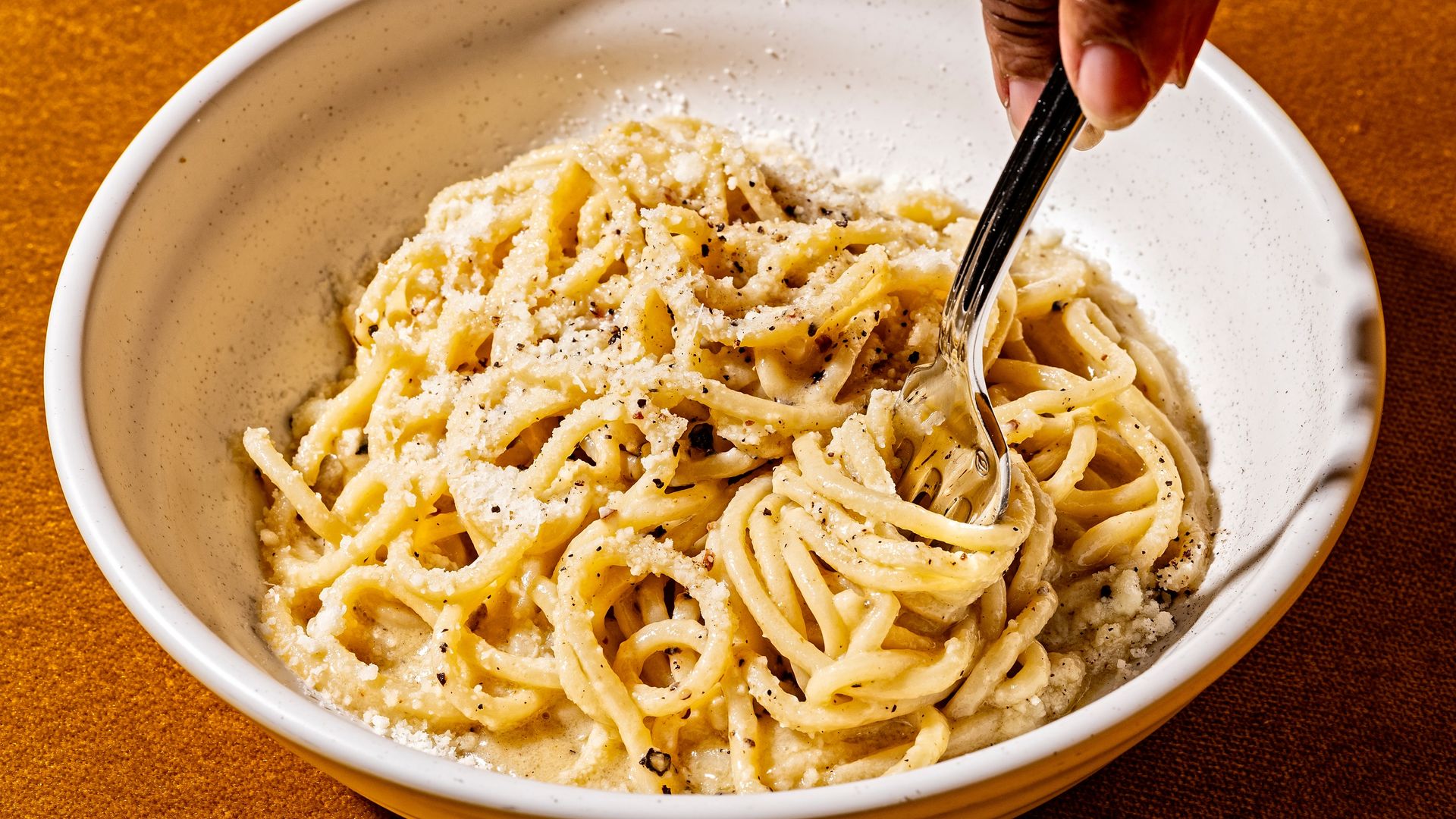 Creamy pasta in a white speckled bowl, sprinkled with grated cheese and black pepper, with a hand twirling noodles using a fork against a brown textured background.