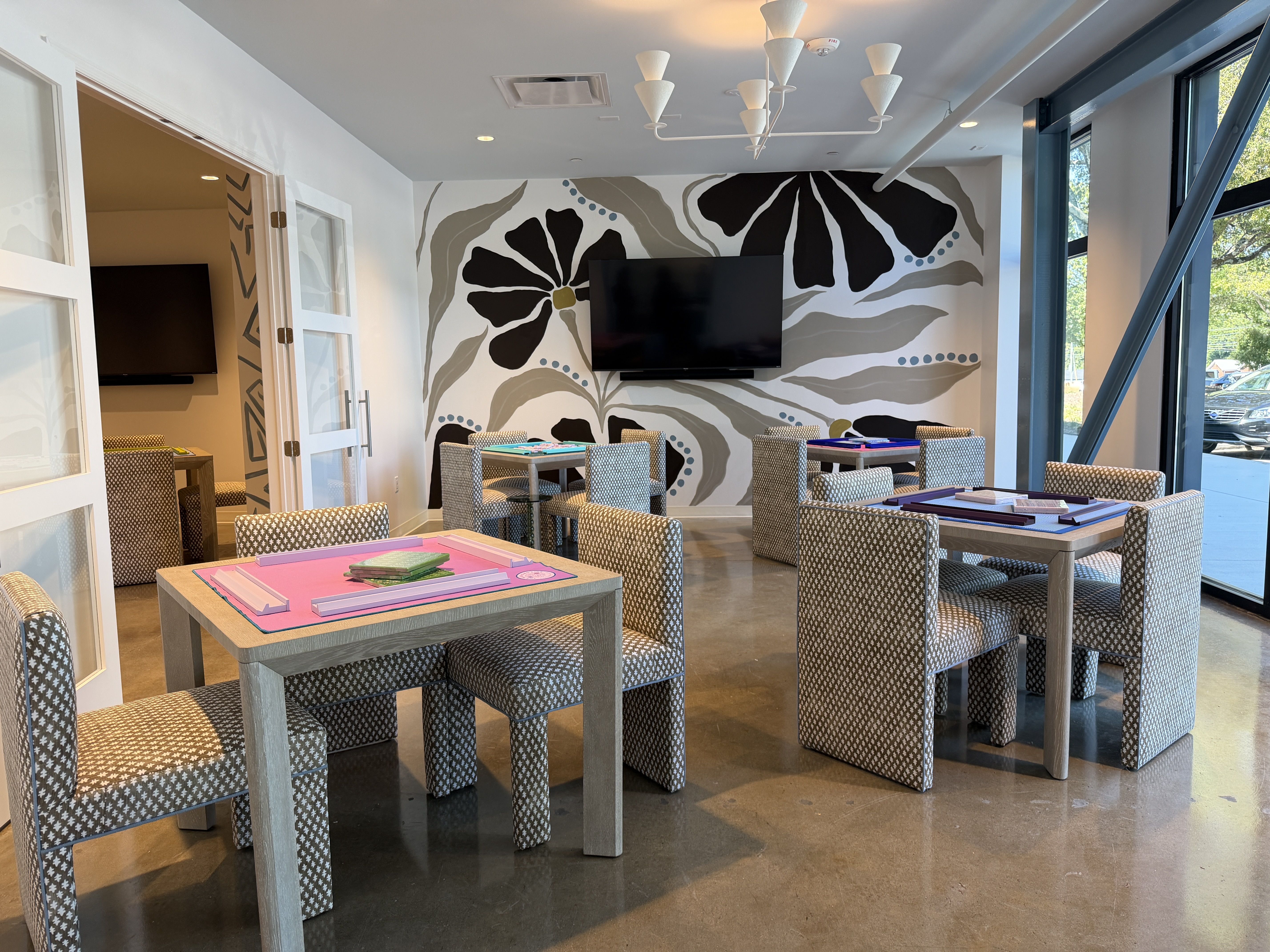 The Queen's Mahjery. Bright modern lounge with gray patterned chairs around light gray tables, a bold black and gray floral mural on the back wall, a wall-mounted TV, and large windows to the right.