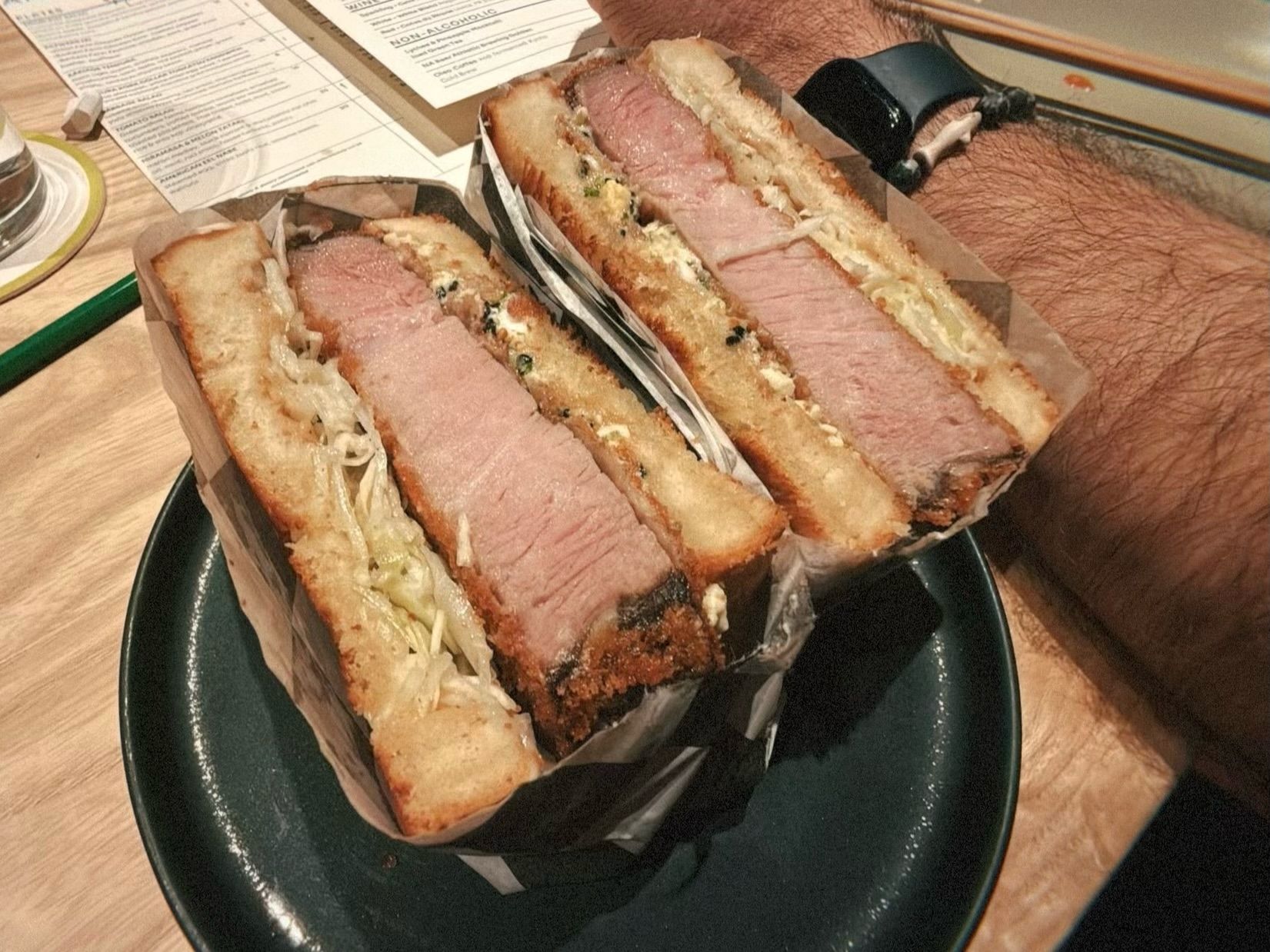 Close-up of a thick-cut ham sandwich with toasted bread, lettuce, and sauce, wrapped in paper on a dark plate. A hairy arm wearing a black wristband is visible beside it.