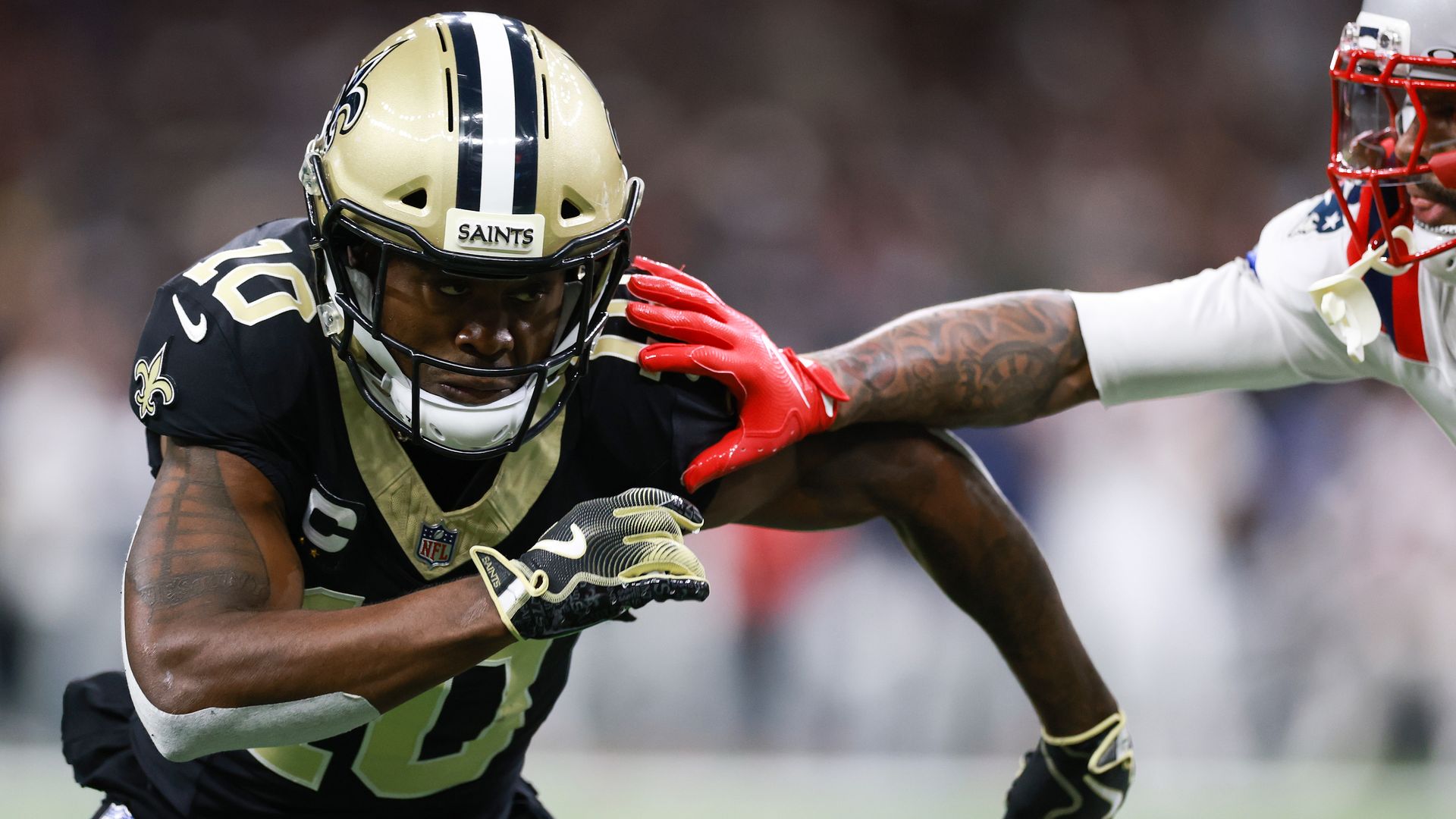 A football player in a Saints uniform runs toward the camera as a Patriots player's arm reaches toward him in an attempt to stop his progress.