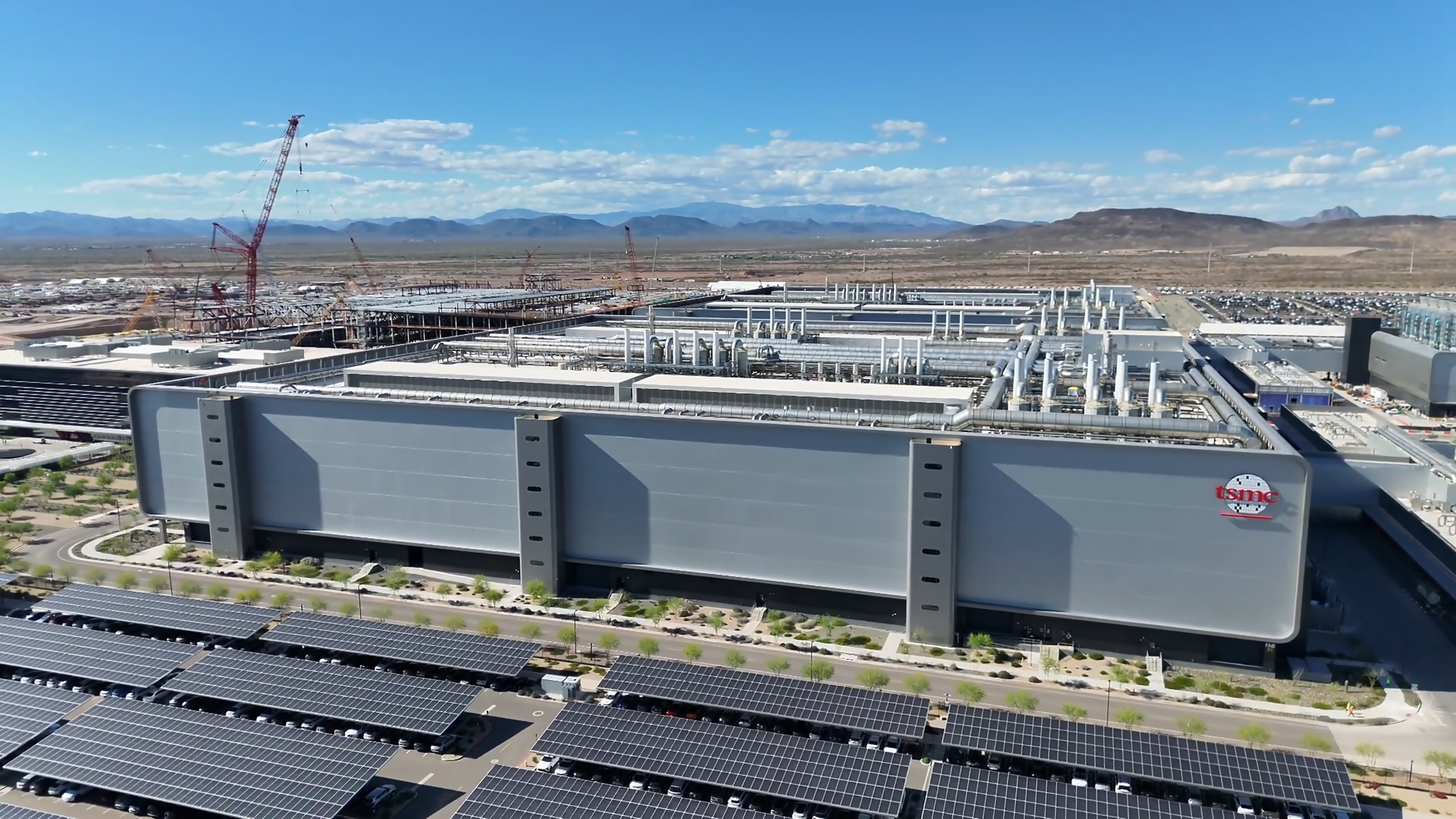 An aerial view of a desert industrial campus with a large gray building, rooftop piping, rows of solar panels in the foreground, and red construction cranes against distant mountains and a blue sky.