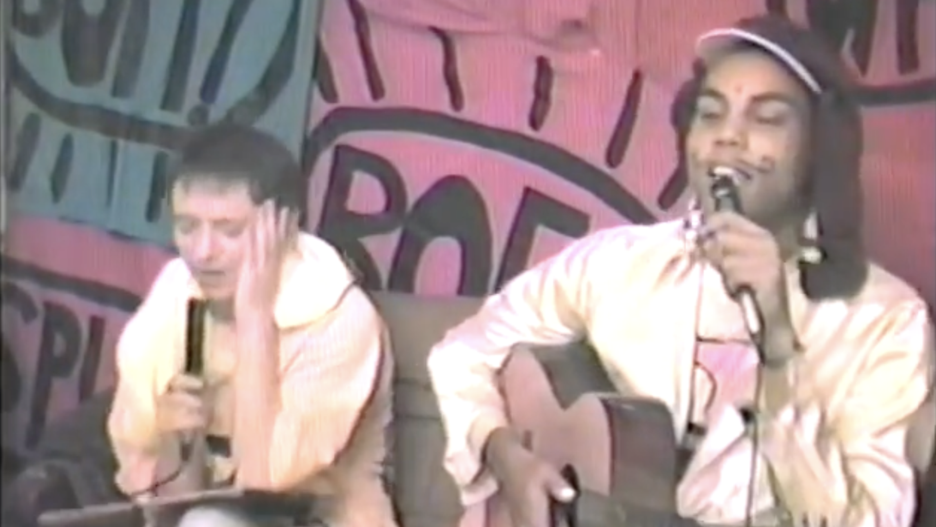 Two men perform on a small stage; one plays an acoustic guitar and sings into a microphone, the other sits beside him with a handheld mic. A pink-and-blue mural backdrop with large lettering behind them.