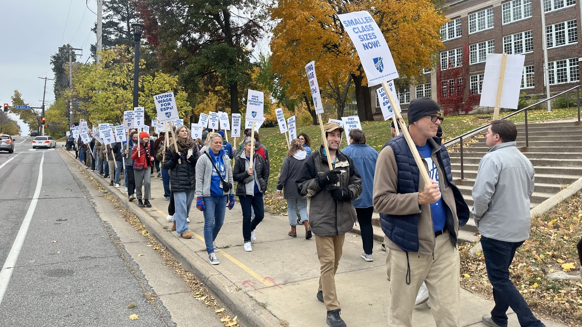 A group of people marching on a sidewalk with signs advocating for smaller class sizes, living wages, pay parity, and special education support, amid autumn trees by a school building.
