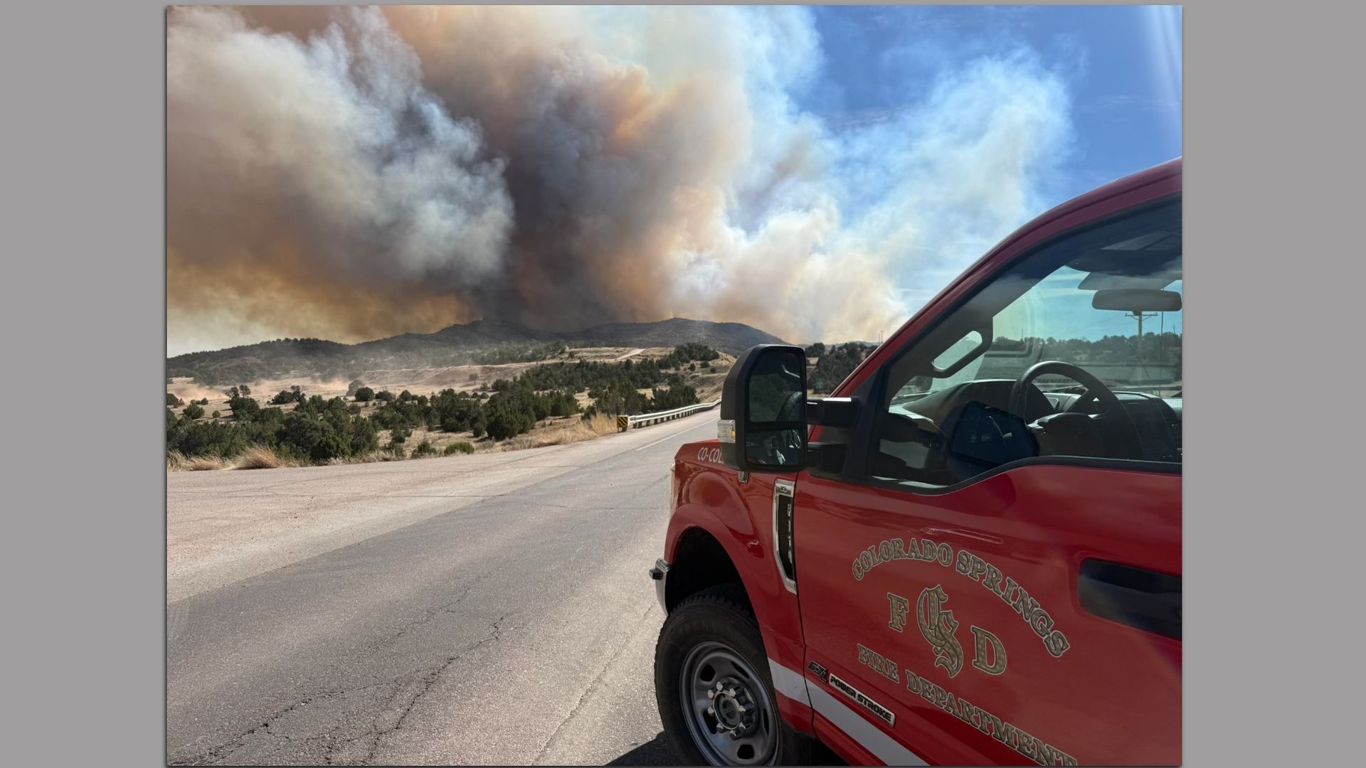 A red Colorado Springs Fire Department truck is parked on a highway at an angle. In the distance wildfire smoke fils the blue sky.