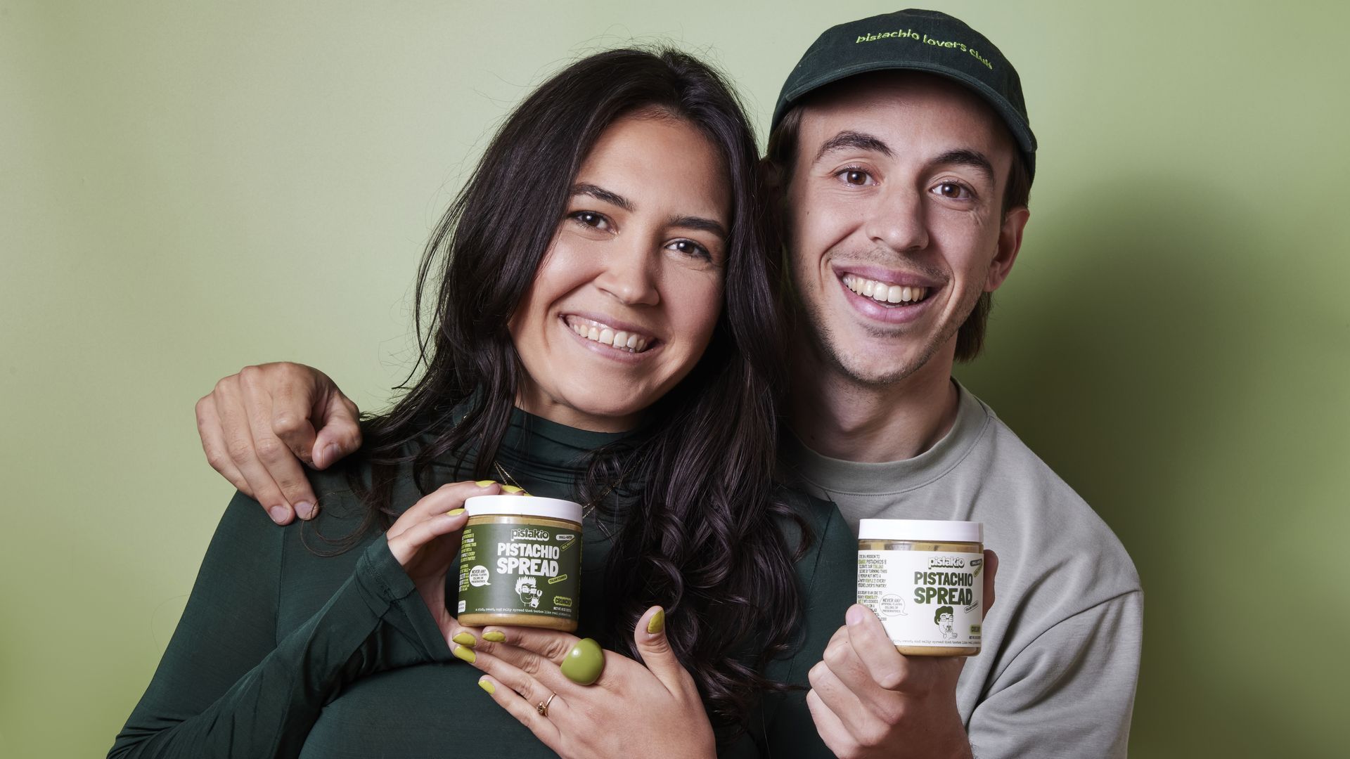 Smiling man and woman with dark hair against green background, holding jars of pistachio spread; woman wears green top, man wears cap with "pistachio lover's club" text.