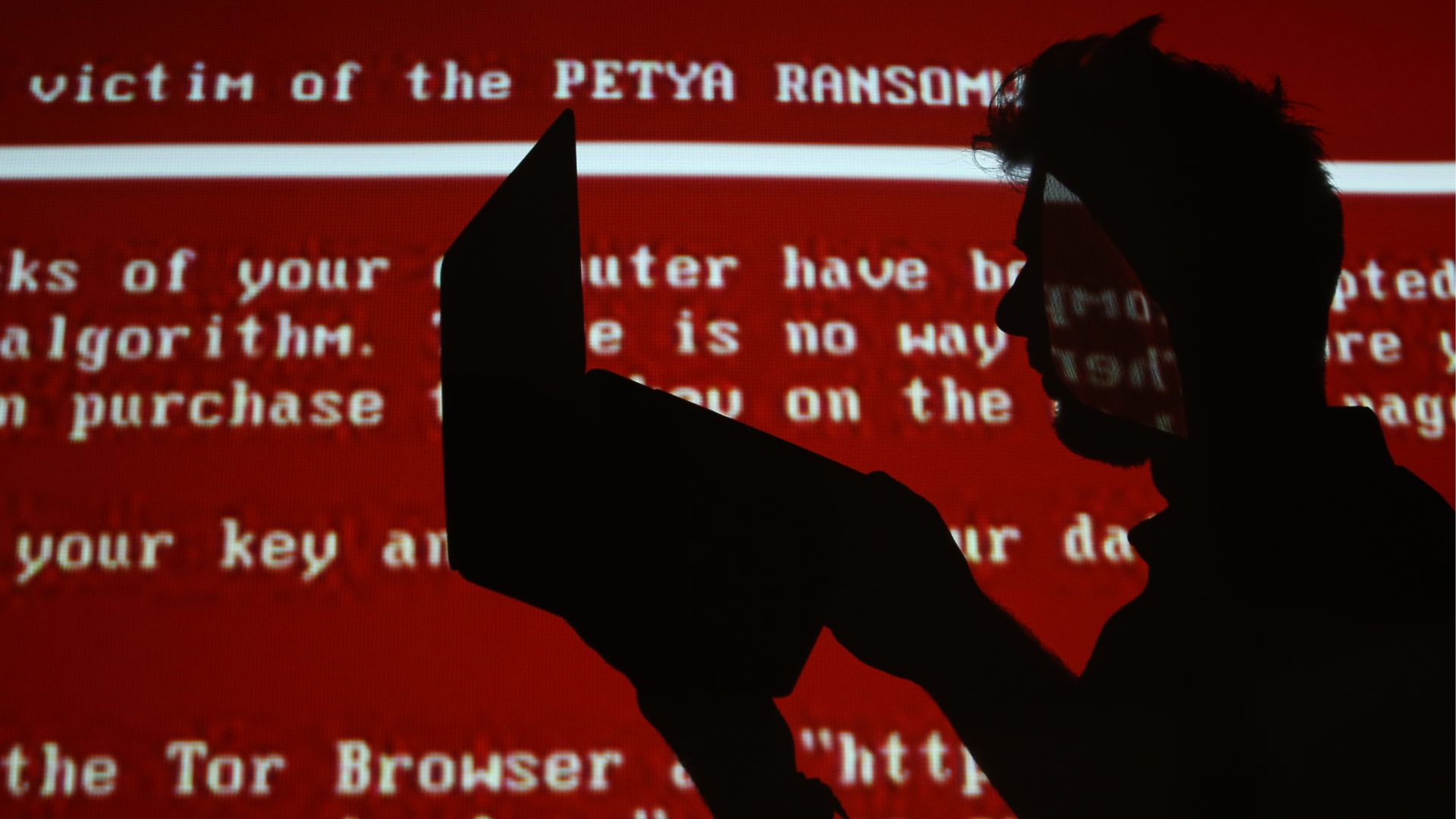Man in silhouette in front of screen projecting NotPetya ransom screen.  