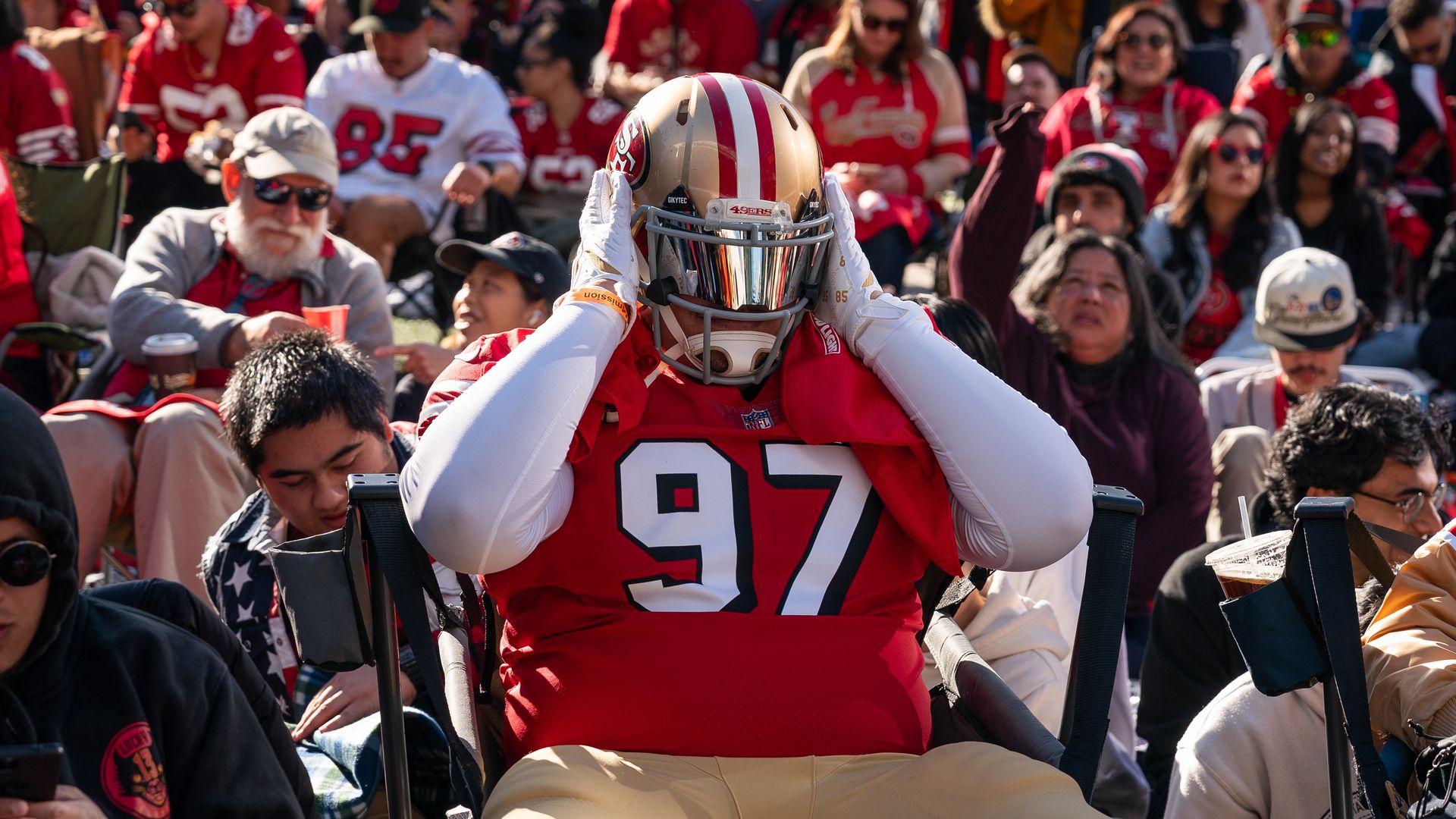 A 49ers fan dressed in the franchise's football gear holds their head as other fans watch the big screen in the back 