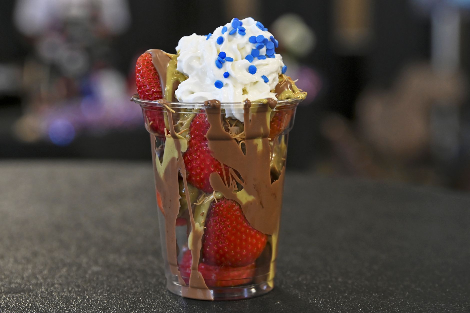 Clear plastic cup filled with fresh strawberries, a drizzle of chocolate, a greenish filling, and whipped cream topped with blue sprinkles.