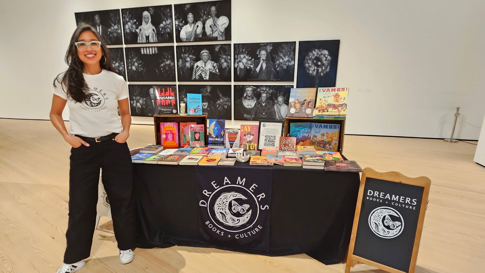Smiling woman with glasses stands next to a book display table labeled "Dreamers Books + Culture" featuring colorful books, inside a light gallery with black-and-white photos on the wall.