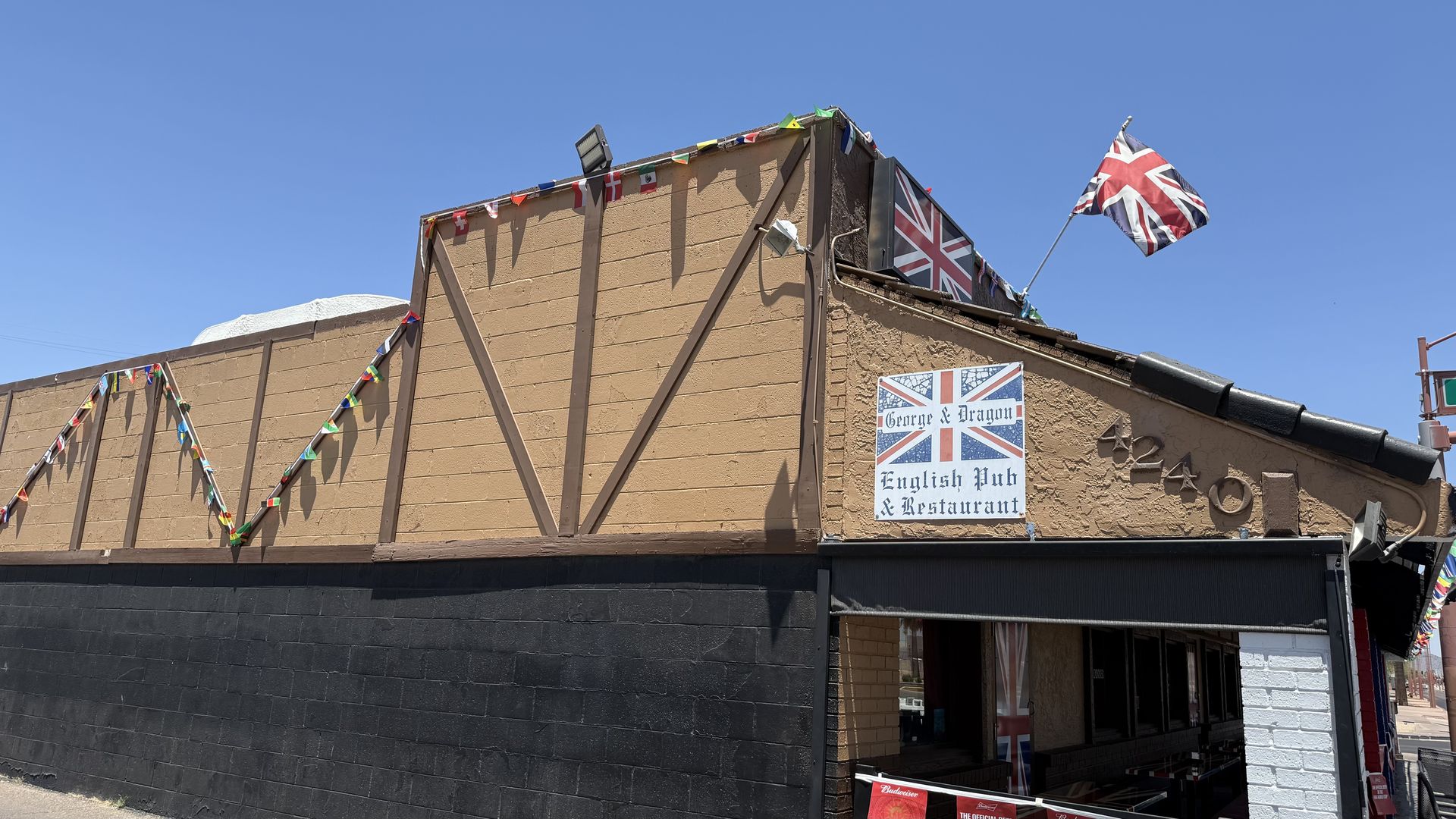 Sunlit brown pub building with a large Union Jack flag on the sloped roof that covers an outdoor seating area, colorful pennant bunting, and a sign reading George & Dragon English Pub & Restaurant on the front.