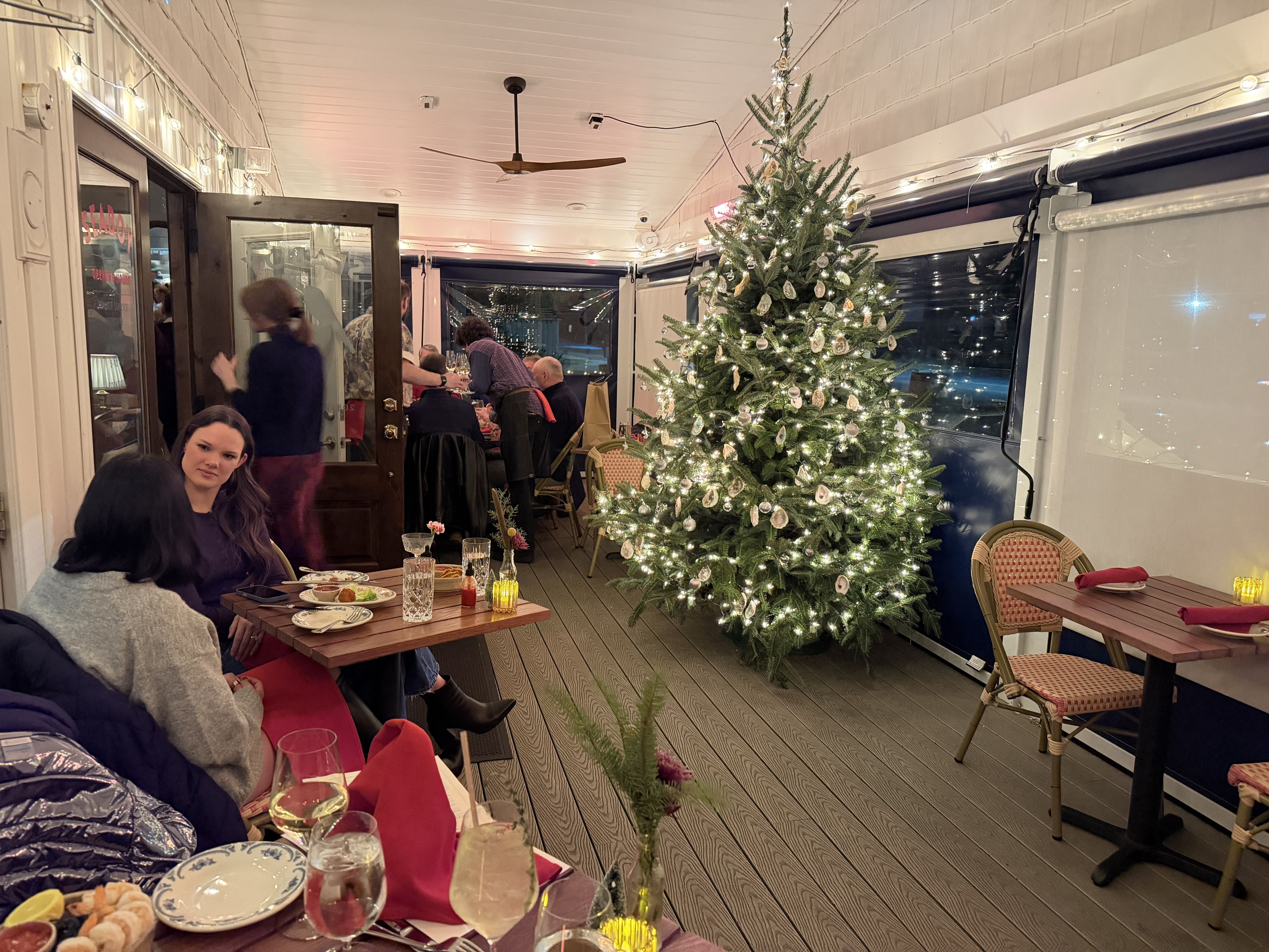 Indoor dining area with wooden floors, people seated at tables with food and drinks, a decorated Christmas tree with white lights and ornaments, string lights on walls, and a ceiling fan.
