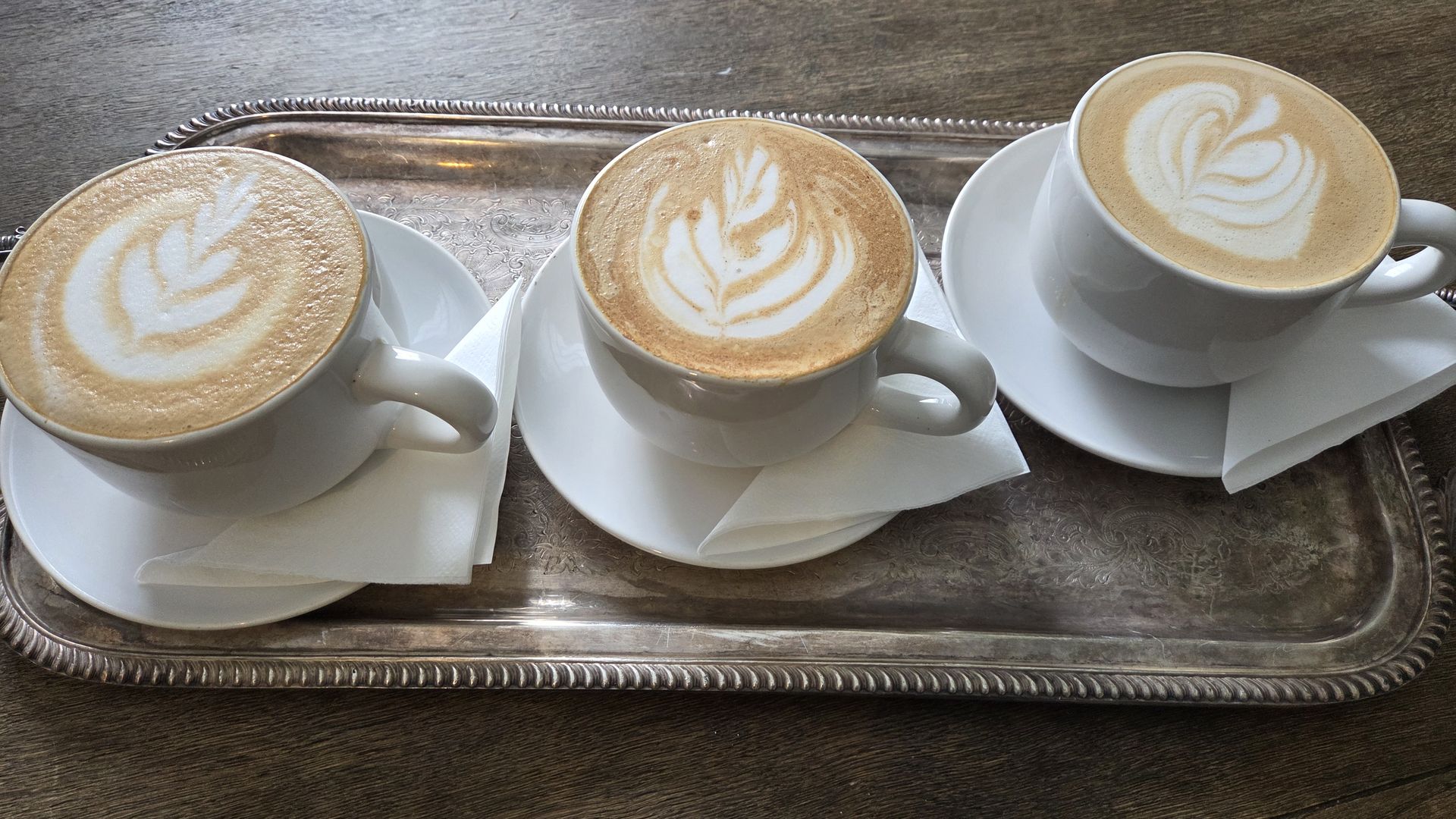 Three white cups of latte with leaf latte art on a silver tray, each cup on a white saucer with a folded white napkin, placed on a wooden table.