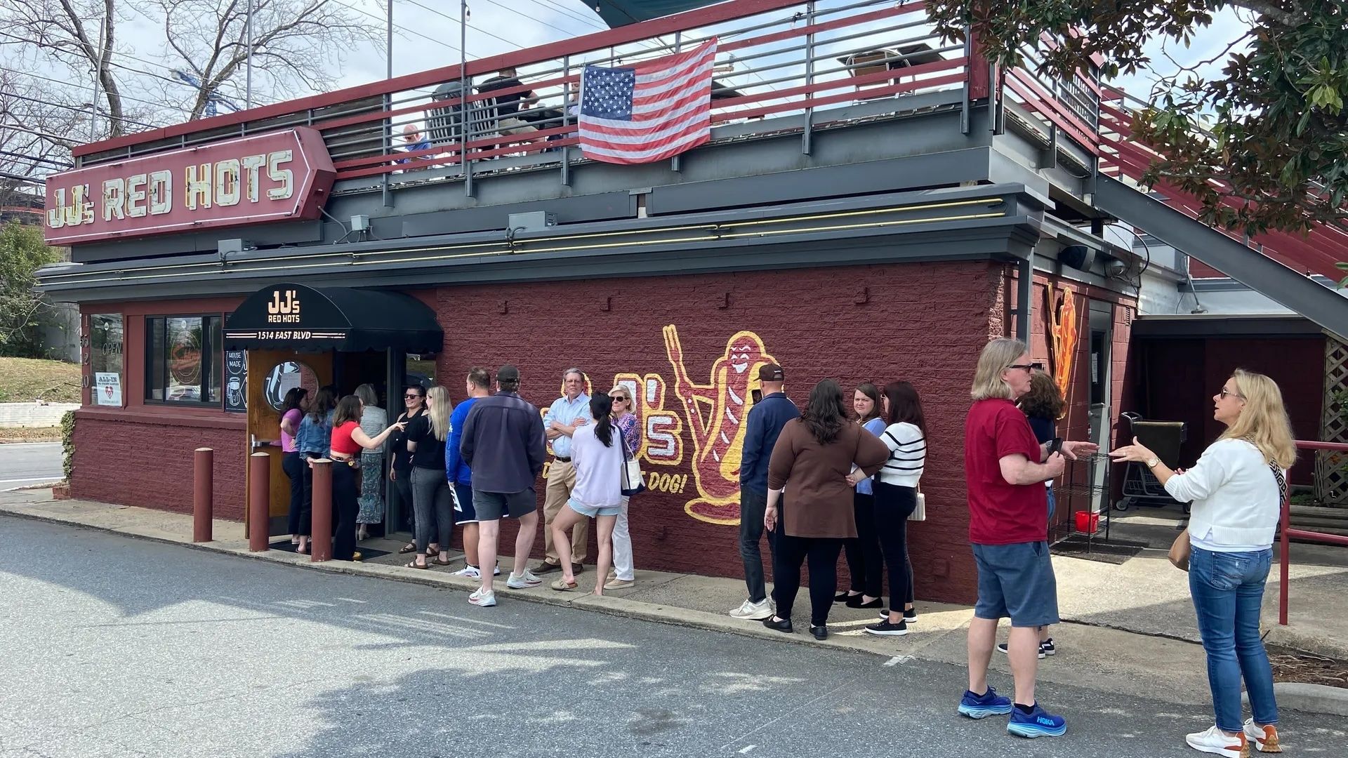 People lined up outside JJ's Red Hots, a red brick building with a large hot dog mural and an American flag hanging from the upper balcony on a clear day.