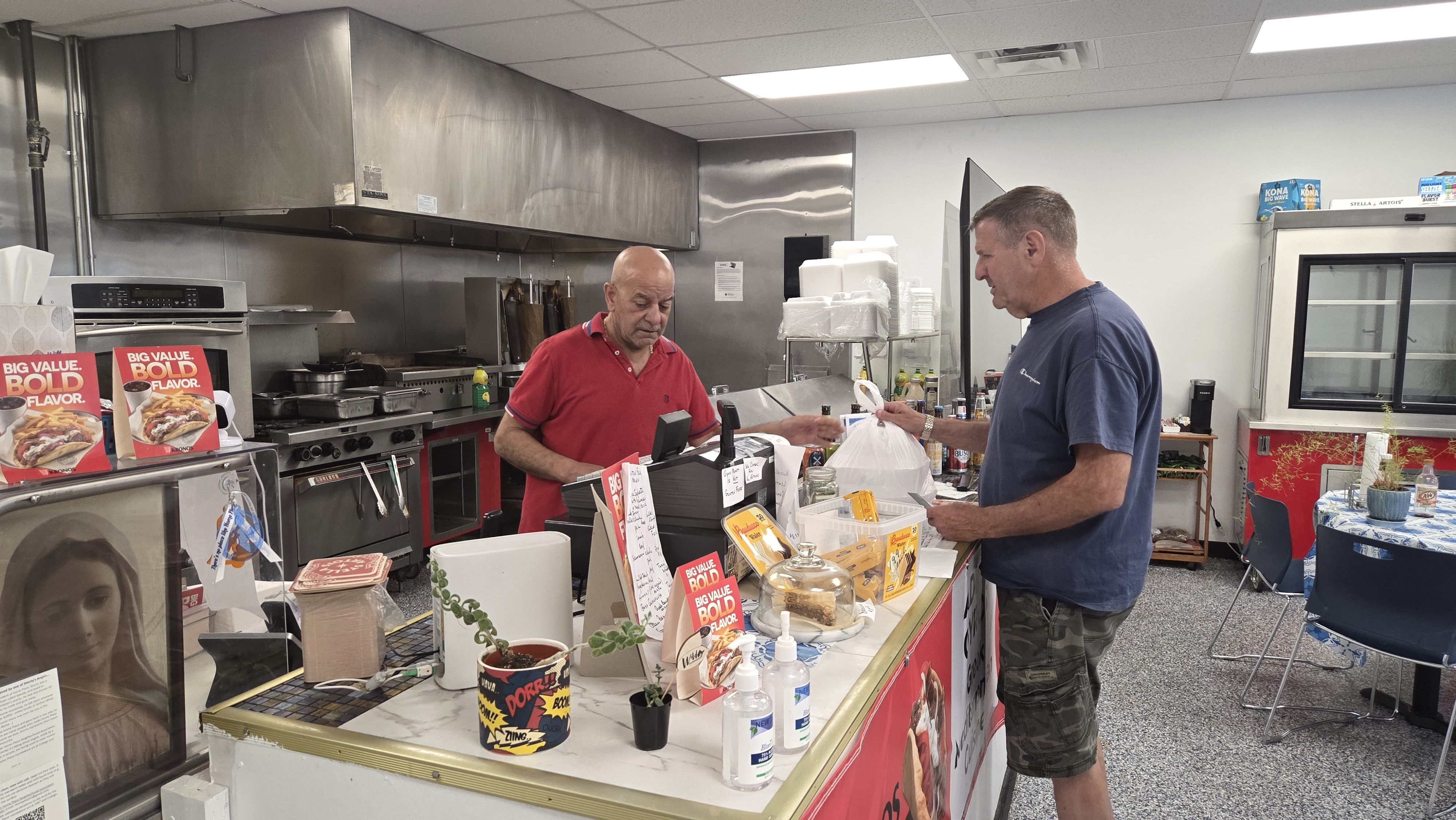 Two men at a deli counter in a bright kitchen. A bald man in a red polo works behind the register while a man in a blue shirt and camo shorts stands at the counter, with stainless steel appliances in back.