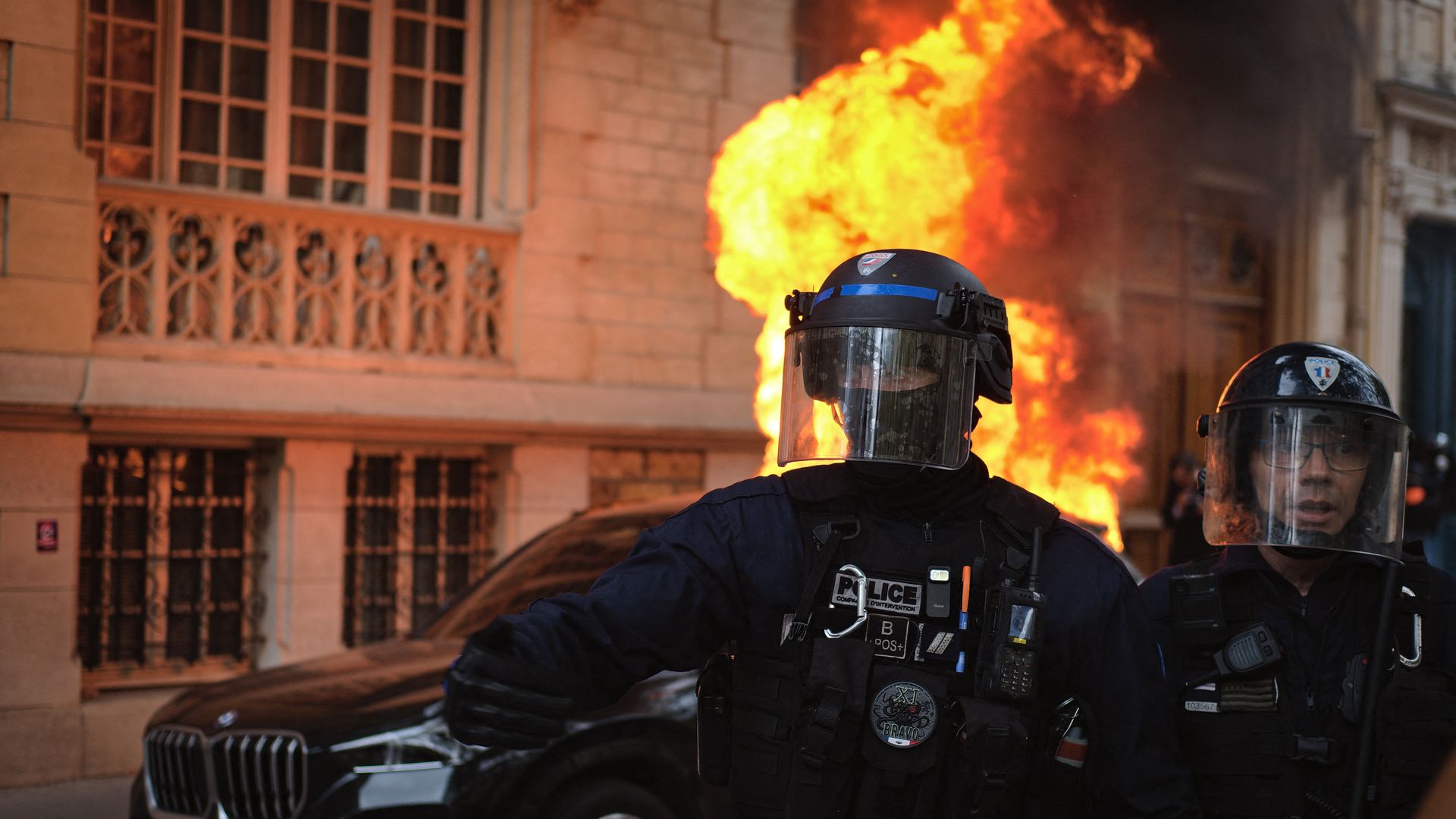 Riot police wearing helmets and protective gear stand in front of a vehicle engulfed in flames during a demonstration in Paris.
