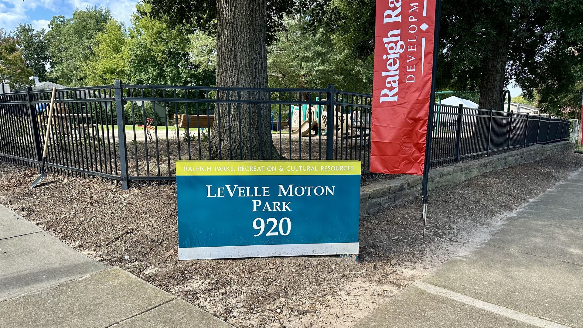 Park entrance with a blue and yellow sign reading LeVelle Moton Park 920, black metal fence, large tree, playground equipment in background, and a red vertical banner for Raleigh Rising Development.