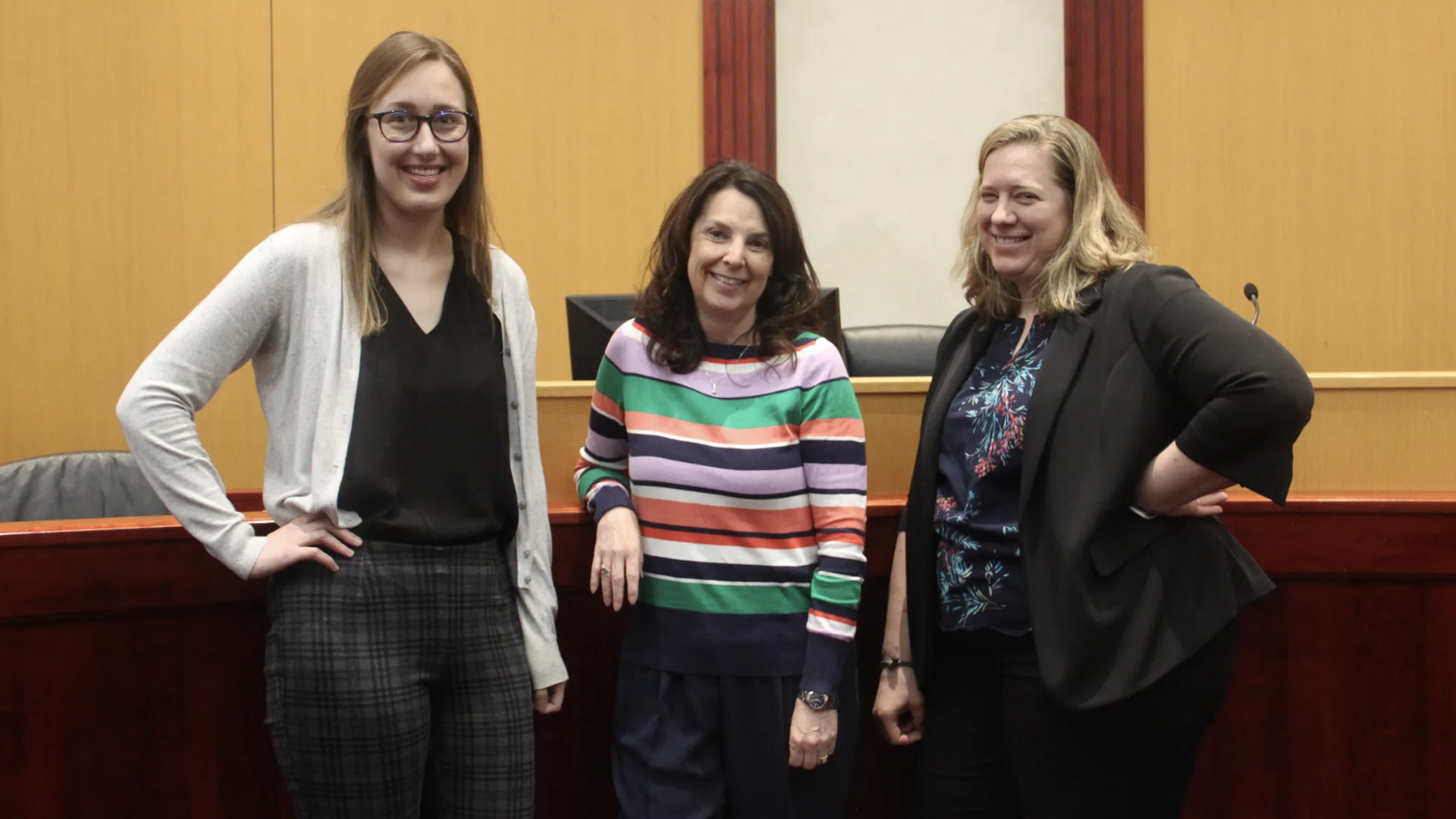Three women stand in a courtroom, smiling. Left: young woman with glasses in a light cardigan and black top; center: woman in a colorful striped sweater; right: woman in a black blazer with a floral blouse.