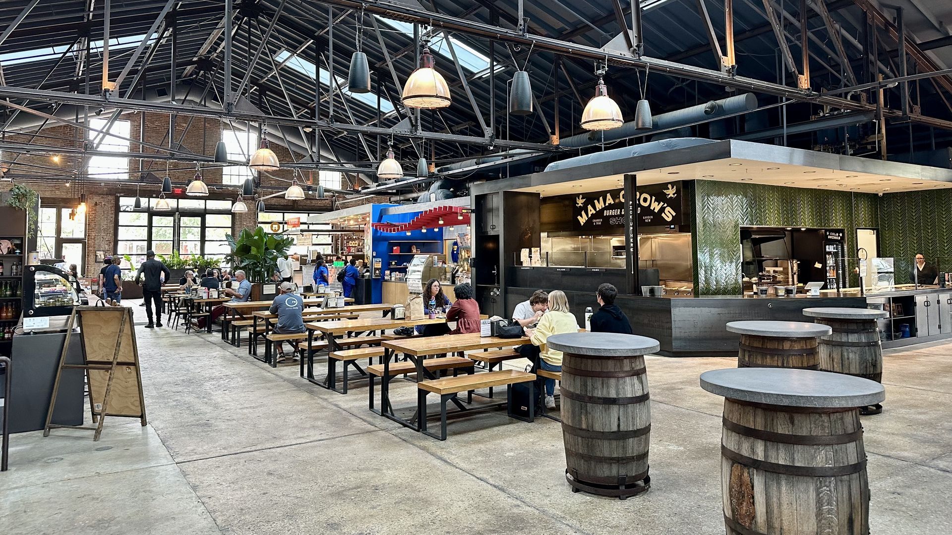 Spacious food hall with high industrial ceiling, wooden picnic tables, and barrel-top tables. People seated and walking near food stalls including one named "Mama Crow's." Bright daylight from skylights.