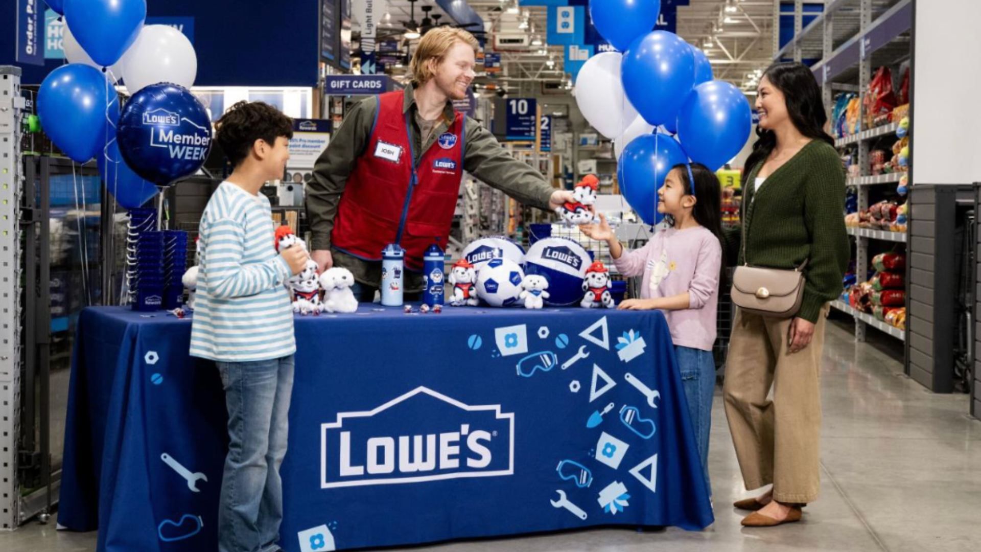 At a Lowe's store event, an employee in a red vest hands a stuffed animal to a smiling girl while her mother watches; blue and white balloons float above a table with Lowe's branding.