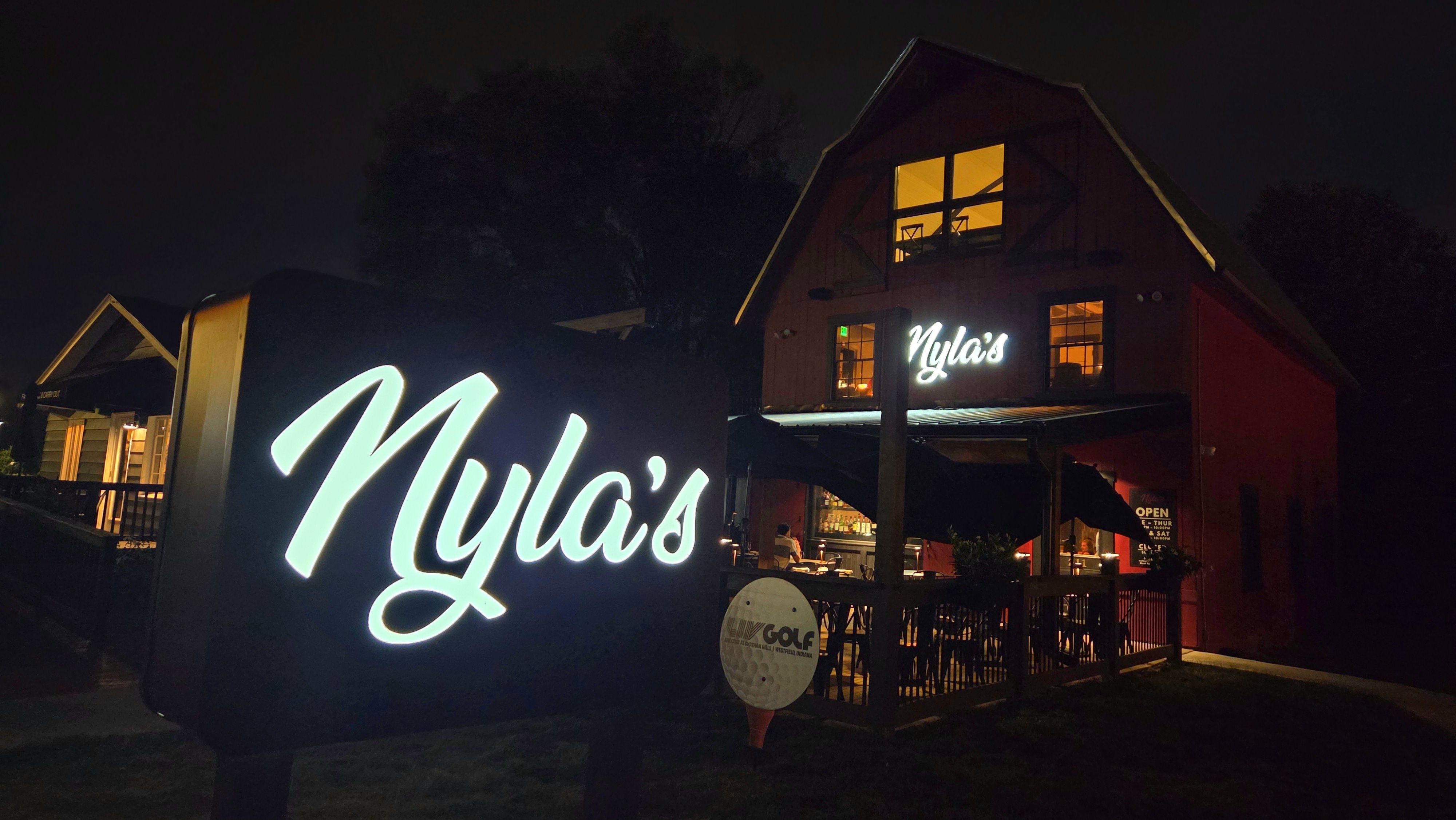 Night view of a red building with lit windows and a bright white neon sign reading "Nyla's". An outdoor patio with tables and umbrellas is visible, along with a large illuminated sign in front.