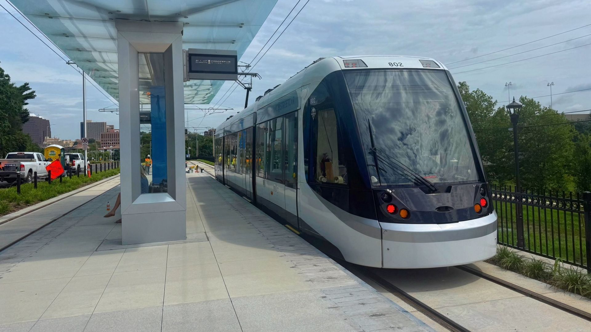 Silver and black modern streetcar at a glass-covered station platform under cloudy sky, with trees and city buildings in the background.