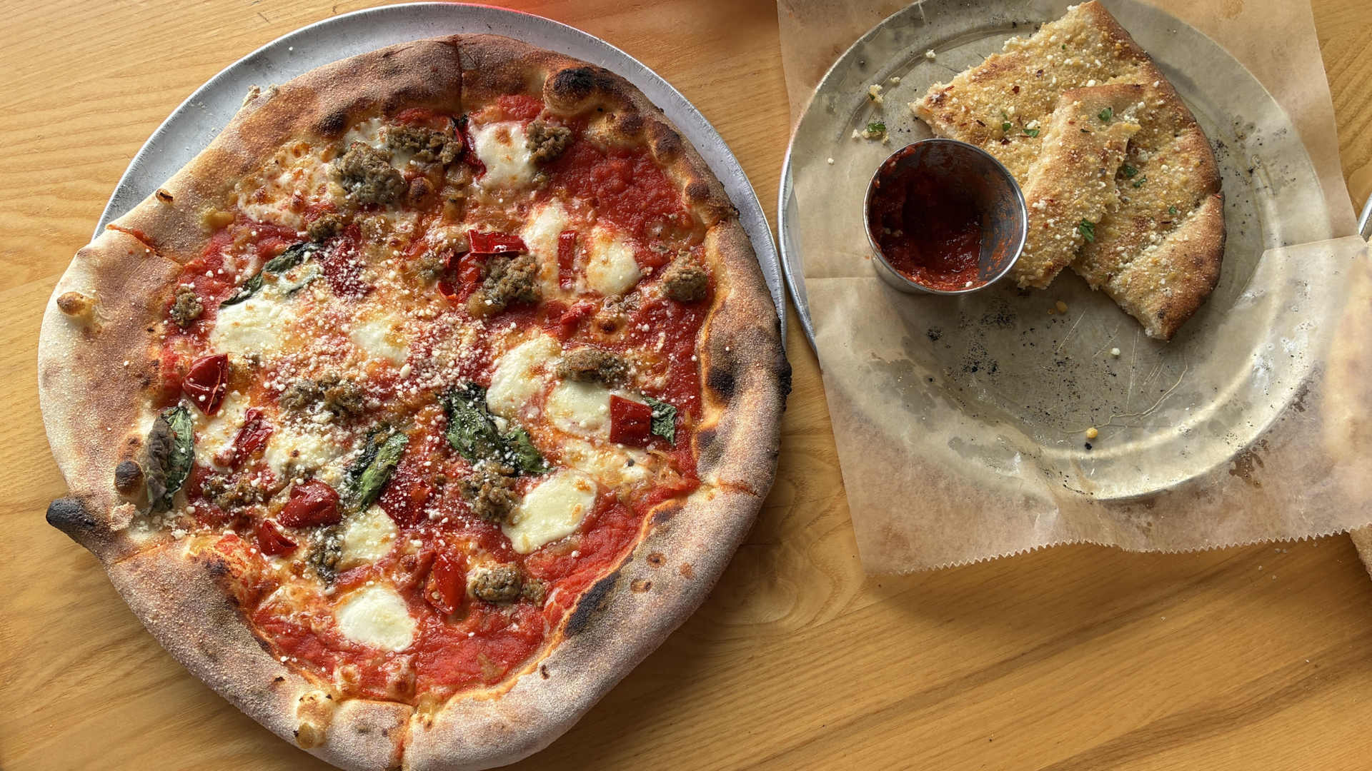 Top-down view of a large pepperoni sausage pizza with red sauce, melted cheese, and basil on a wooden table. Beside it, a plate of garlic bread slices with a metal cup of marinara.