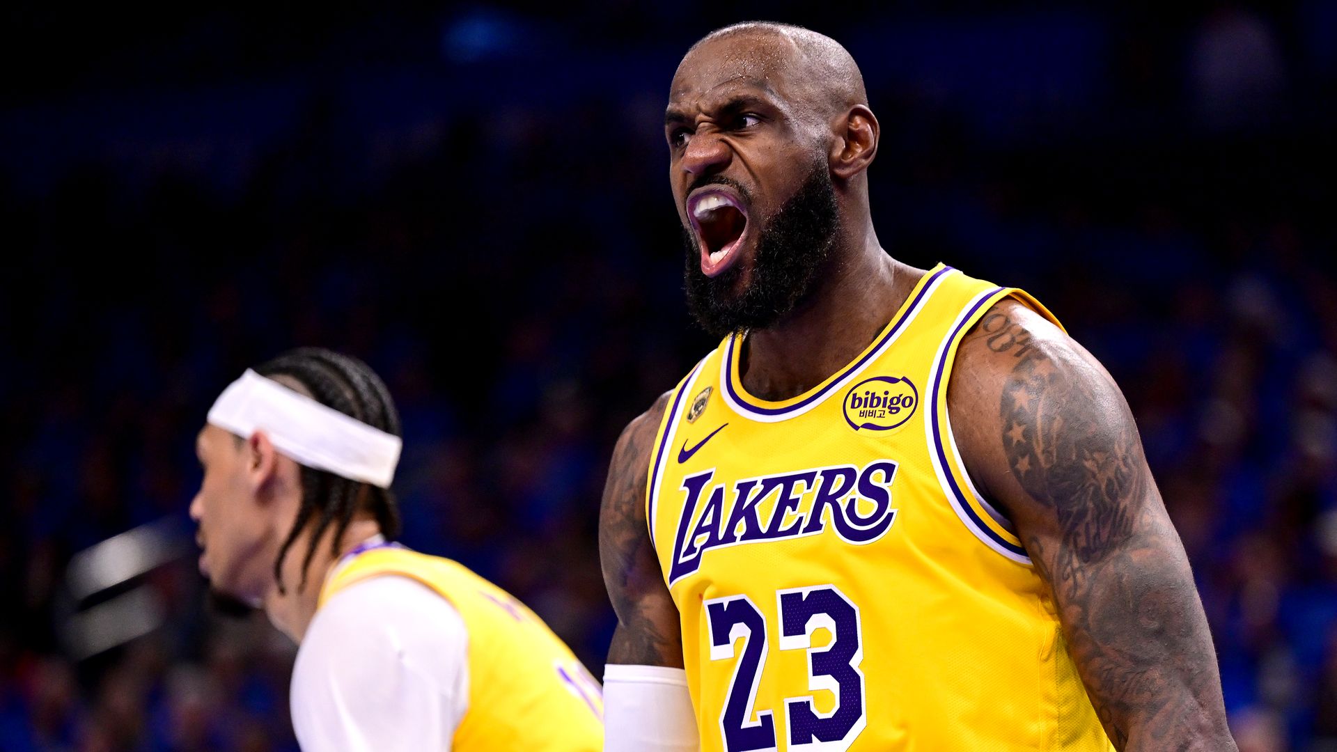 Close-up of a basketball player in a yellow Los Angeles Lakers jersey (#23) with purple trim, shouting with mouth wide open in a dark arena; teammate with a white headband blurred in the background.