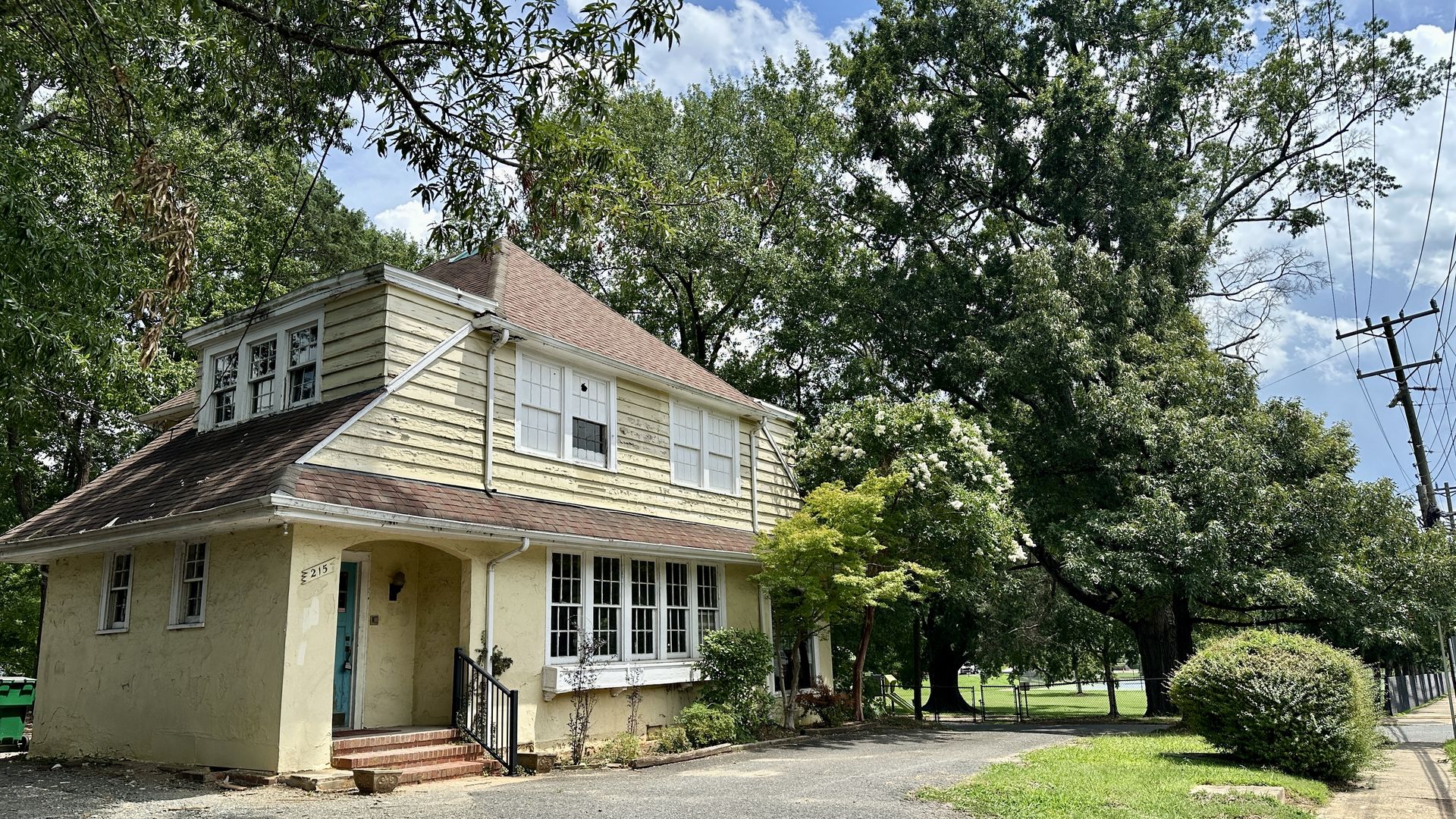 crescent park elizabeth. charlotte. Sunny view of a pale yellow two-story house with white trim, red roof, and many leafy trees surrounding it. Blue sky, scattered clouds; power lines on the right and a driveway in front.
