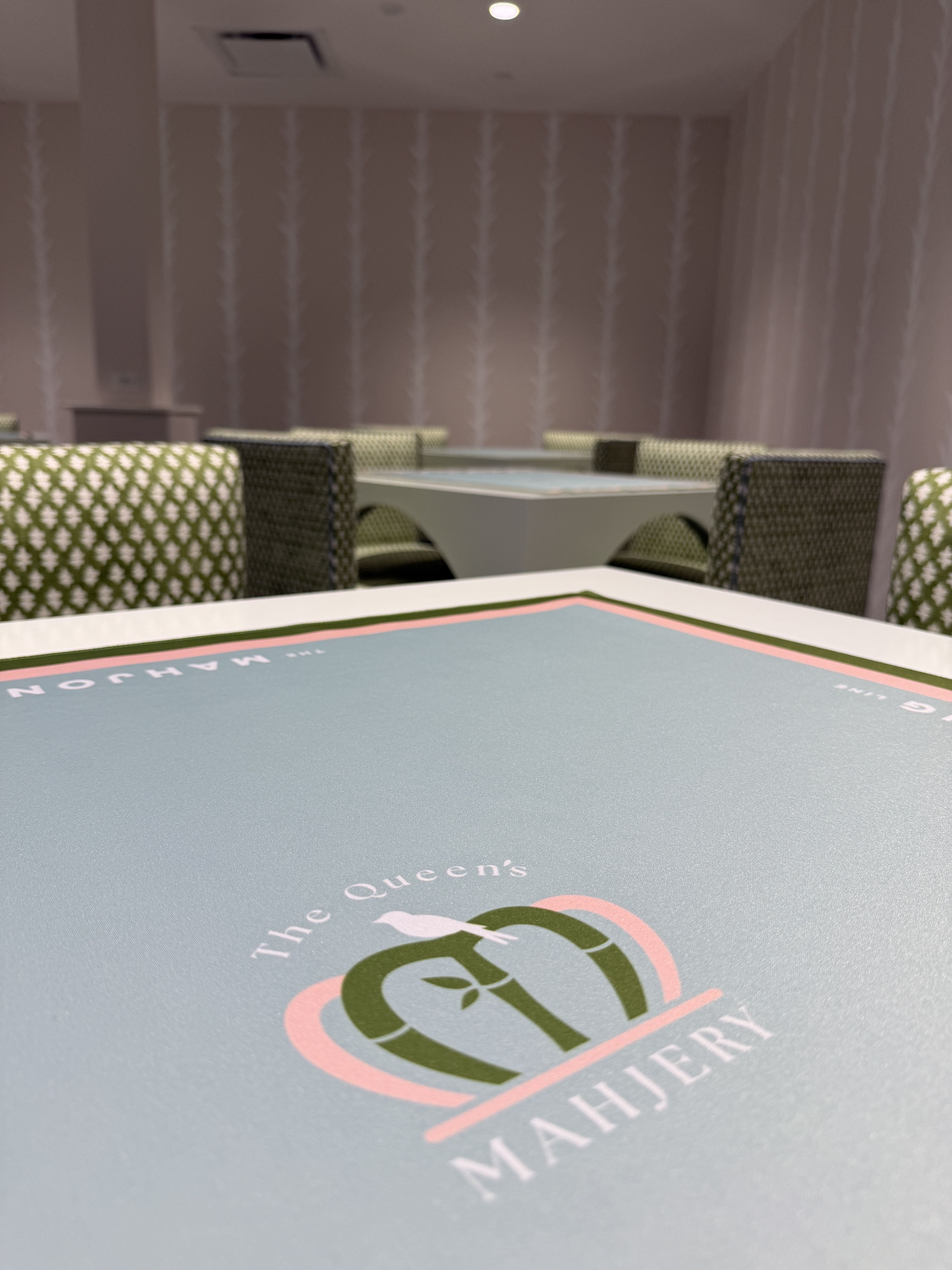 The Queen's Mahjery. Low-angle view of a game room: a blue mahjong table in the foreground, green patterned chairs around white tables, and beige walls with vertical stripe wallpaper; soft ceiling light overhead.