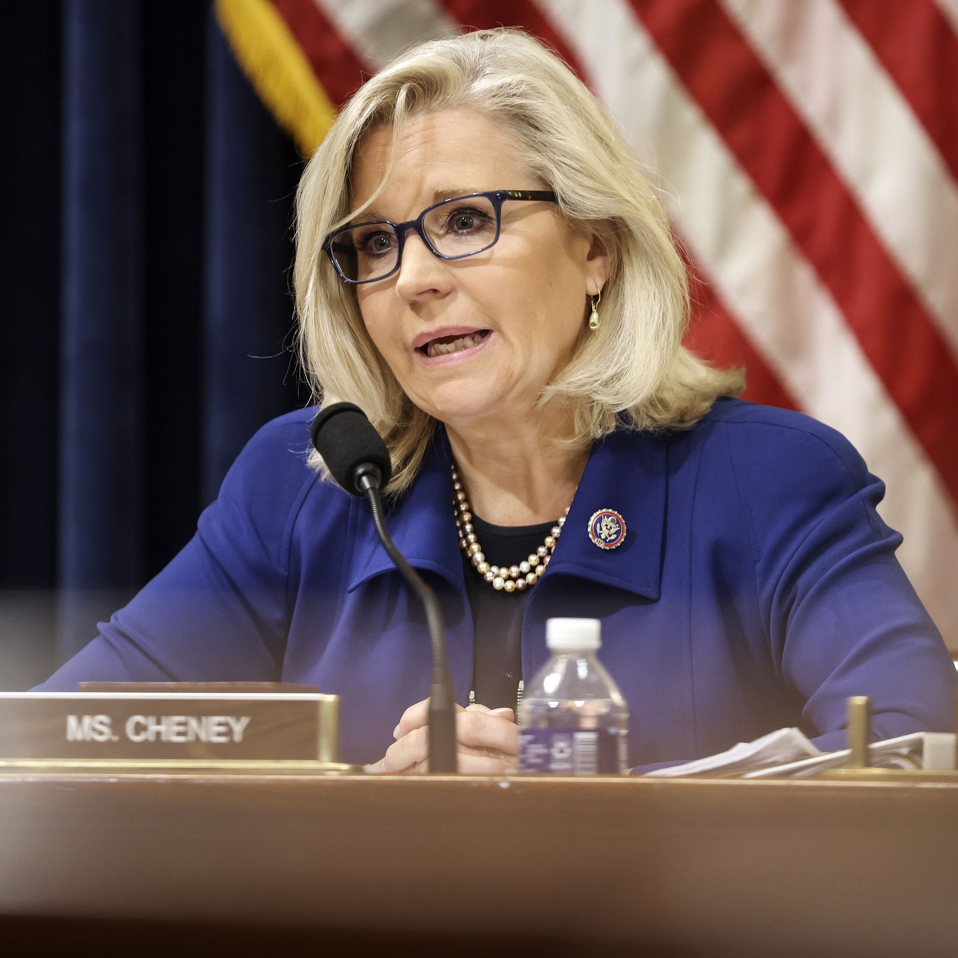 Rep. Liz Cheney (R-Wyo.) in a blue blazer sits at a table, behind a microphone and a placard of her name, and in front of an American flag.