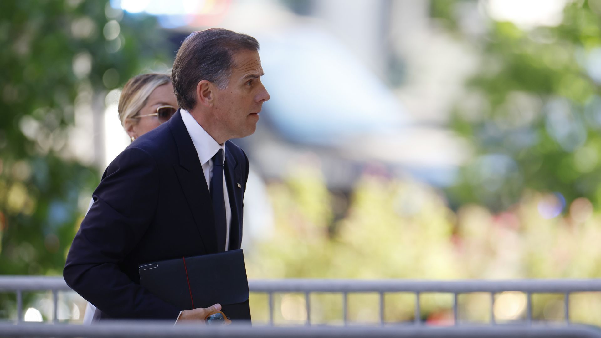 Hunter Biden walks outdoors through a gate. He is wearing a dark suit and tie with a white shirt. 