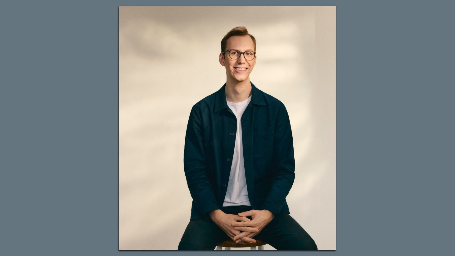 Portrait of a smiling young man with glasses and light brown hair, wearing a dark teal jacket over a white shirt, seated on a stool against a warm beige studio backdrop.