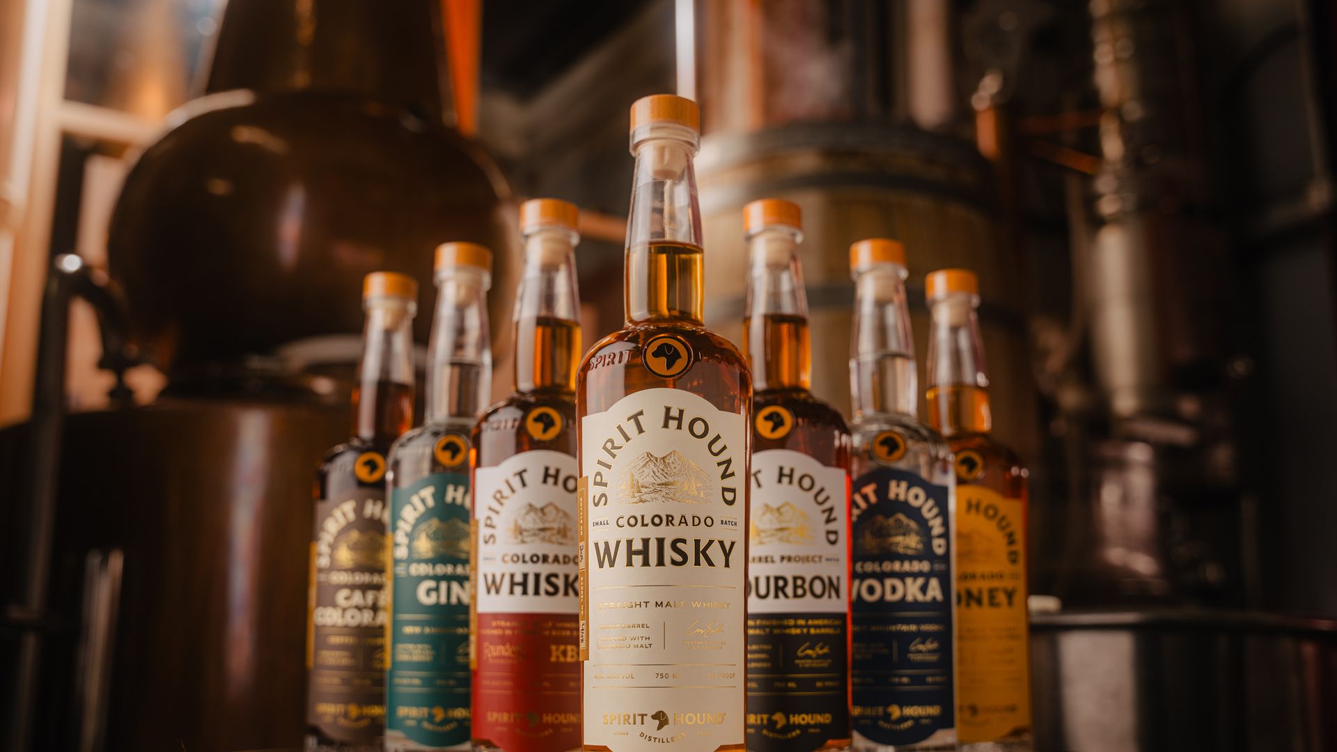 Seven Spirit Hound liquor bottles in varied colors on a wooden barrel in a dimly lit distillery. Center bottle is Colorado Whisky with amber liquid, surrounded by gin, bourbon, vodka, and honey labels.