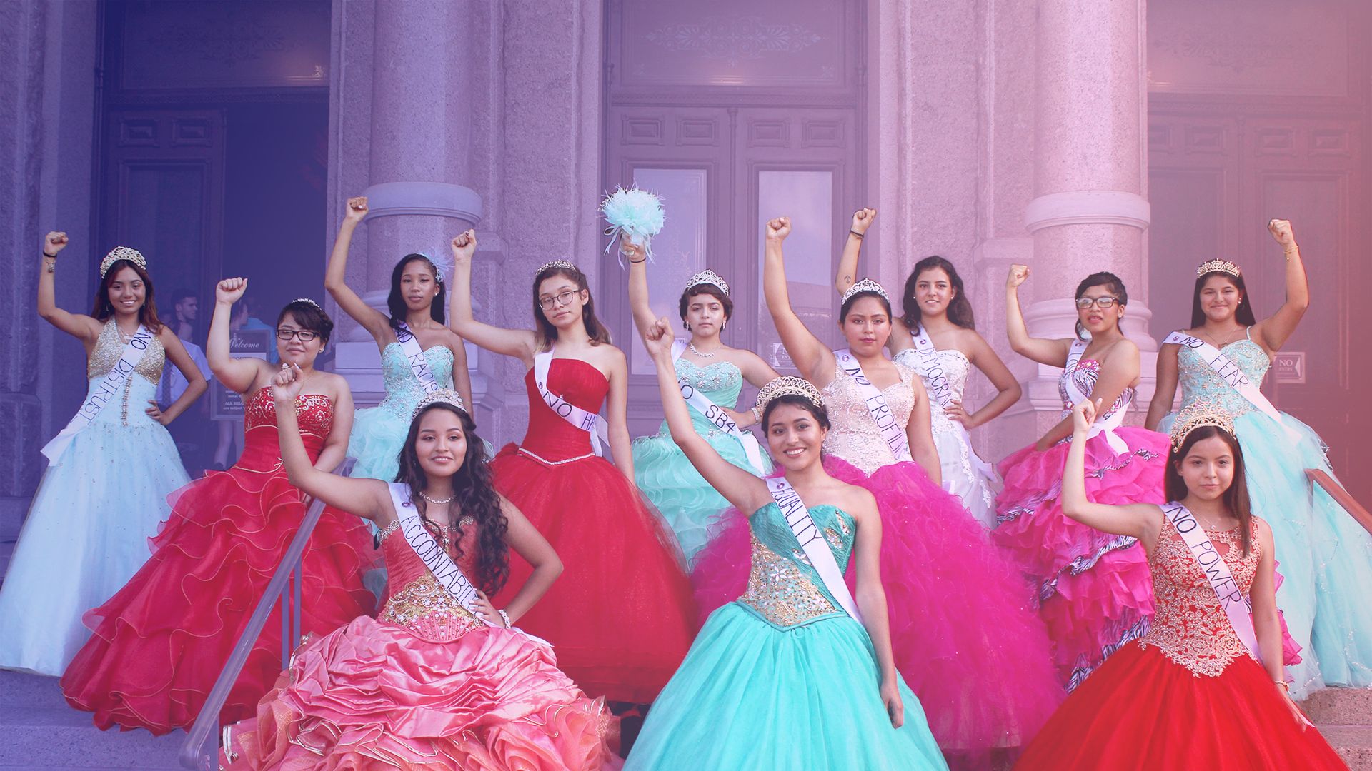 A group of young women in quinceanera dresses stand with their fists in the air