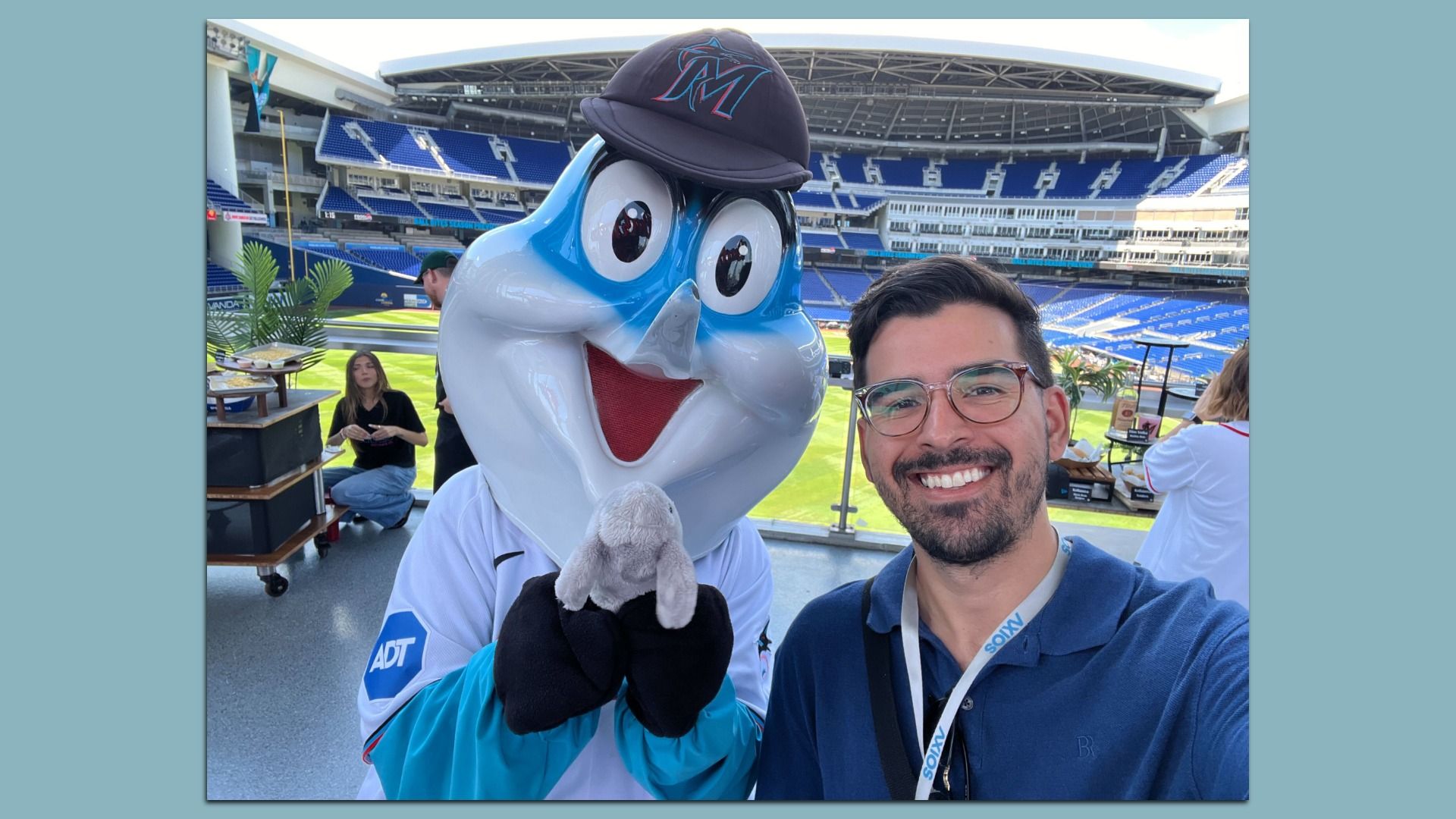Selfie with a large blue-and-white cartoon mascot wearing a black cap at a baseball stadium. A smiling man with glasses stands beside it on the field, with blue seats and green grass in the background.