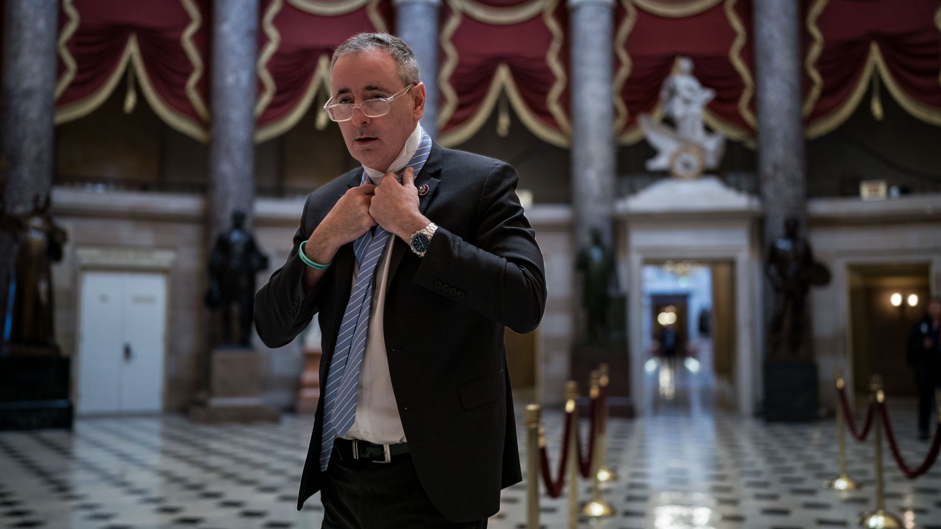 Rep. Brian Fitzpatrick, wearing a dark gray jacket, white shirt and clear glasses, tying a light blue tie as he walks through statuary hall in the U.S. Capitol.