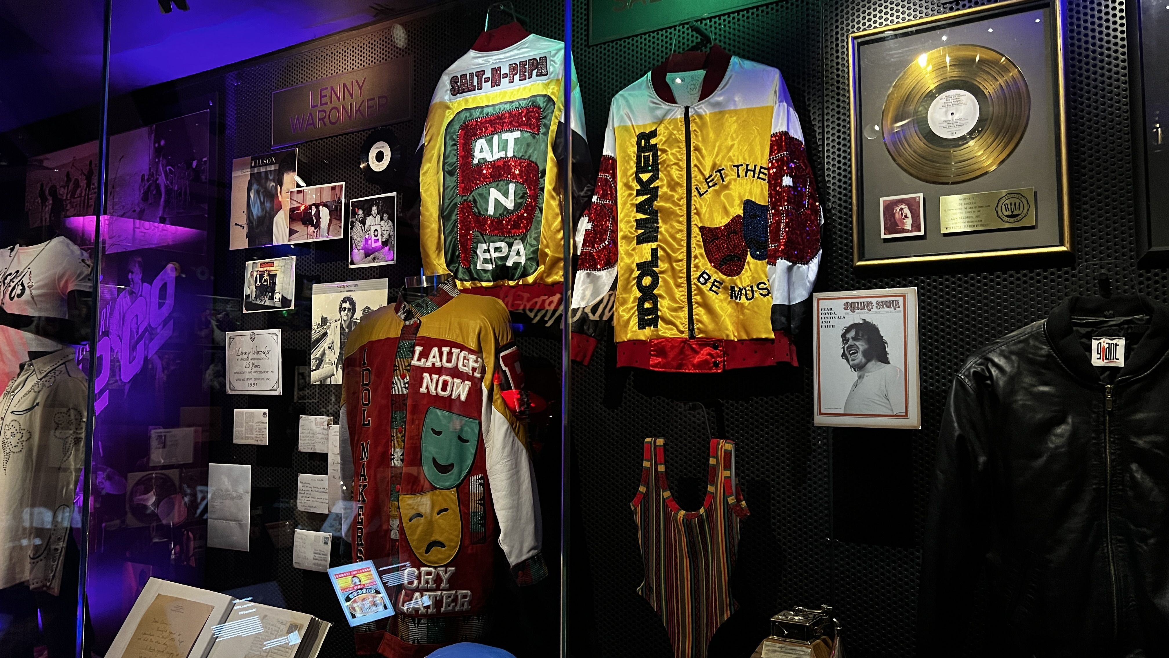 Display case with colorful jackets, including Salt-N-Pepa and Billy Idol-themed designs, a striped tank top, photos, documents, a gold record, and a black leather jacket in a museum exhibit.