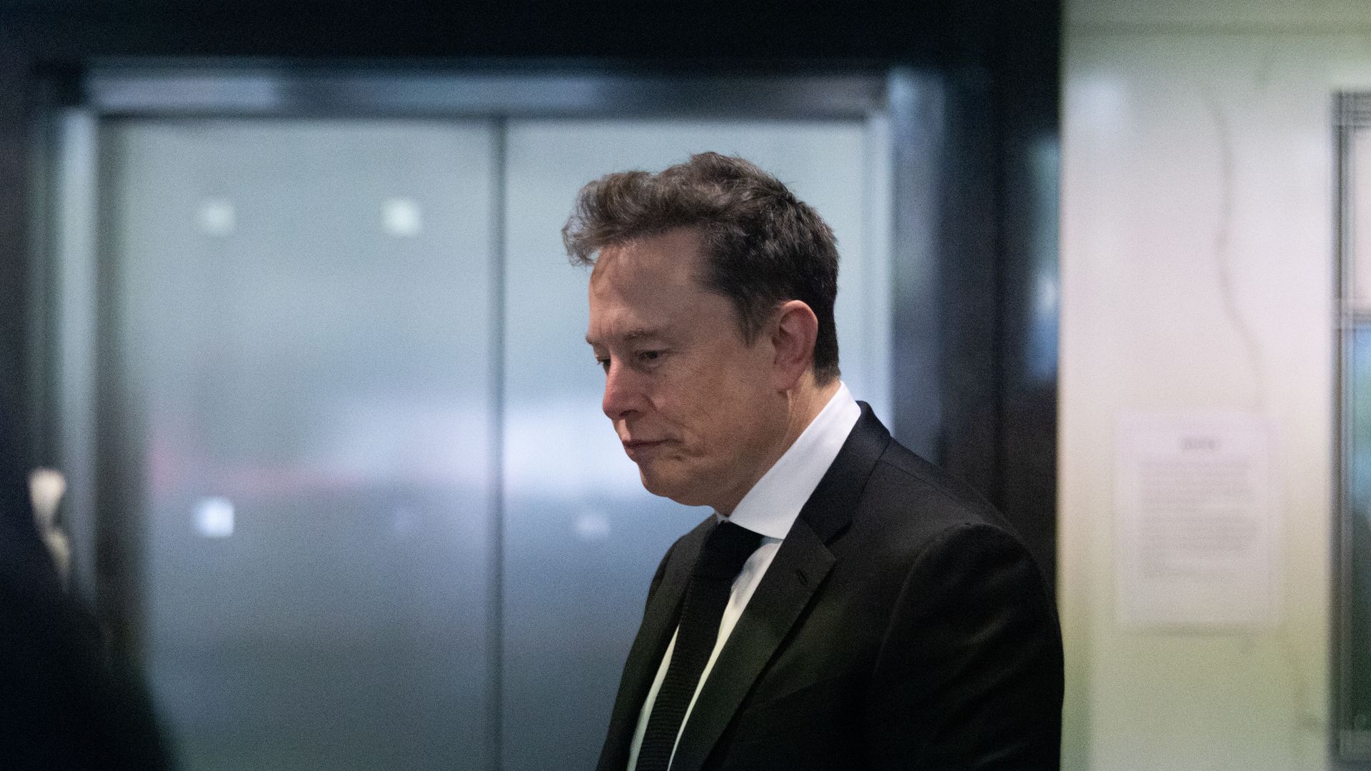 Side view of a man in a dark suit, white shirt, and black tie, looking down. He stands in a modern interior near glass doors and pale walls.
