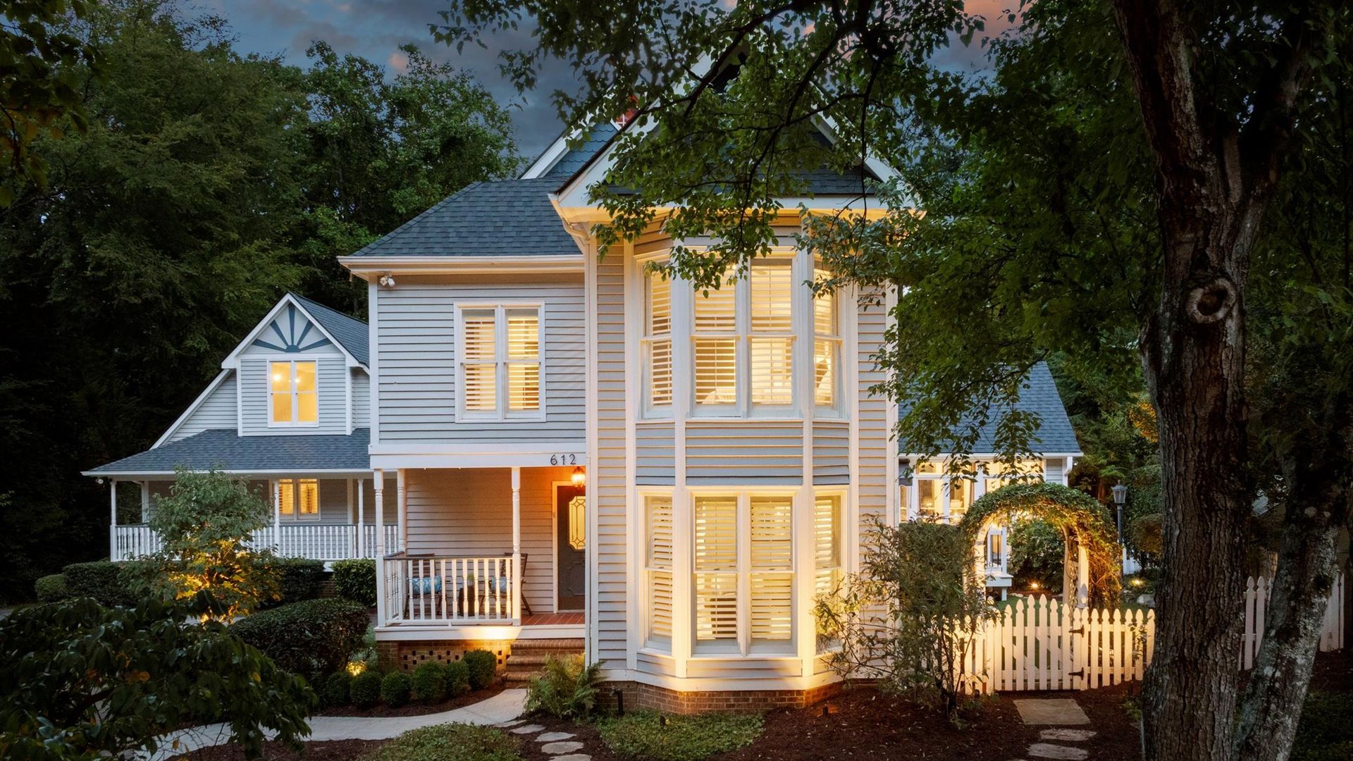 Two-story gray house with white trim and lit windows at dusk, surrounded by greenery, a stone path, a white picket fence, and a tree in the foreground under a cloudy sky.