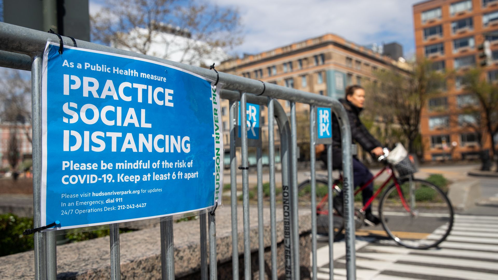 Photo of a blue sign with white text that says "Practice social distancing" hung on a fence in a metropolitan area