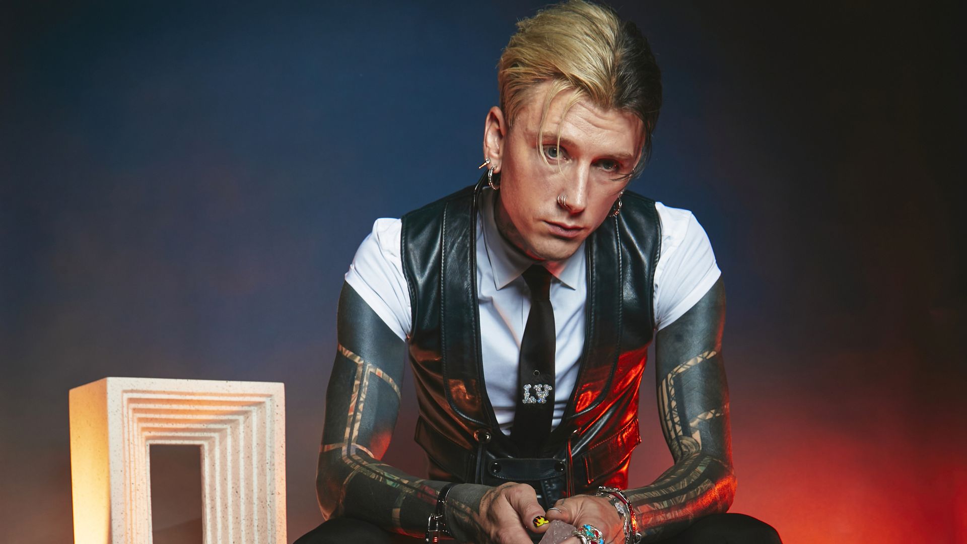Machine Gun Kelly poses for a photo with slicked back hair and a black vest. 